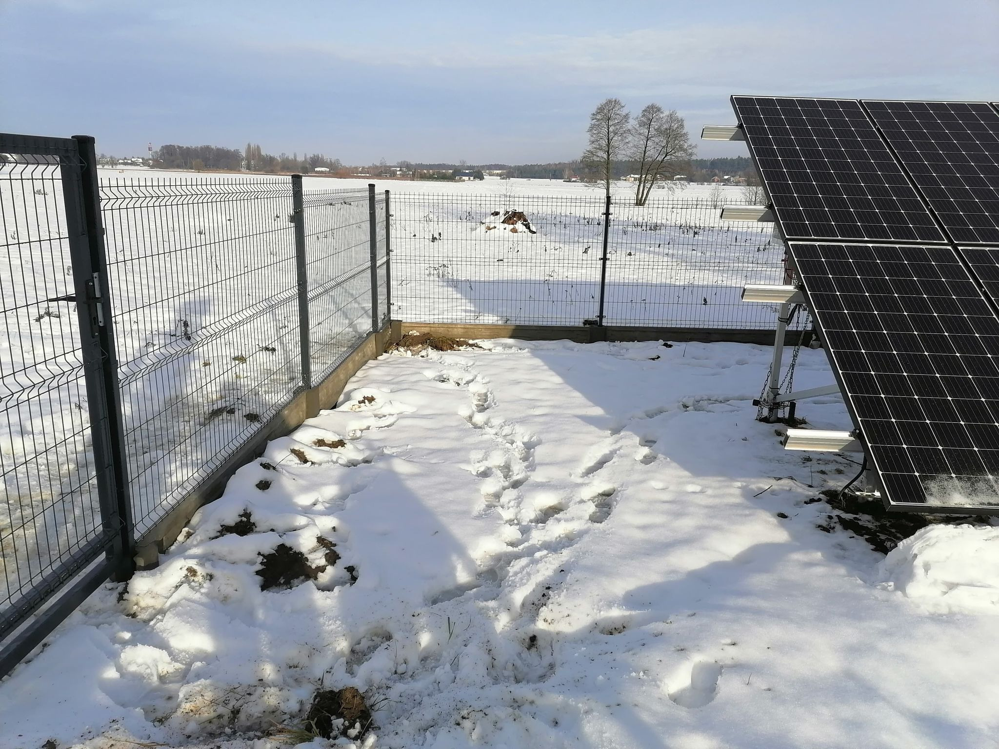 Szare ogrodzenie panelowe z widocznymi śladami stóp w śniegu, obok panele słoneczne, w tle zimowy krajobraz z drzewami i zabudowaniami.