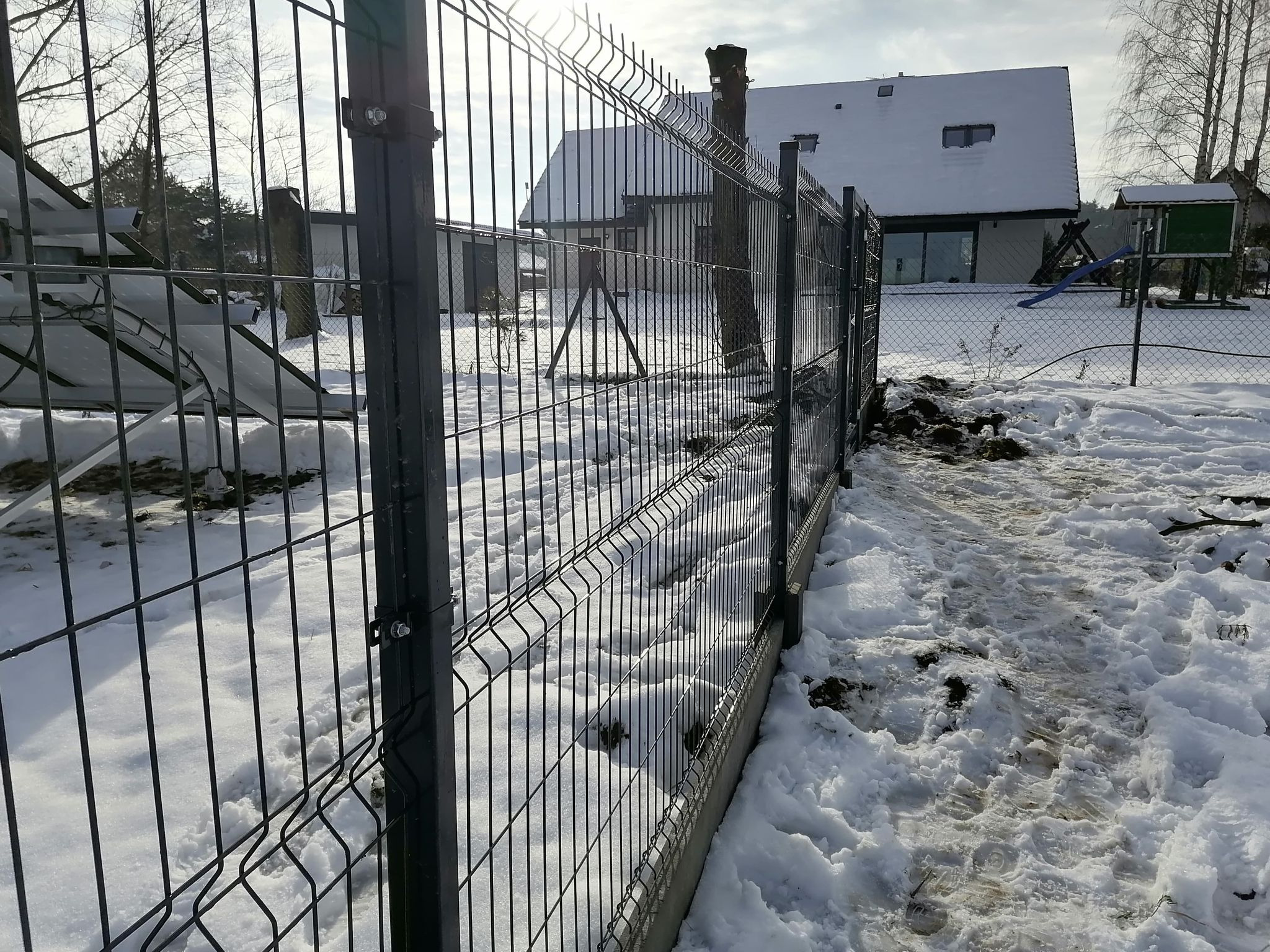 Nowoczesne, ciemne ogrodzenie z paneli stalowych na tle zasypanego śniegiem podwórka z domem i placem zabaw