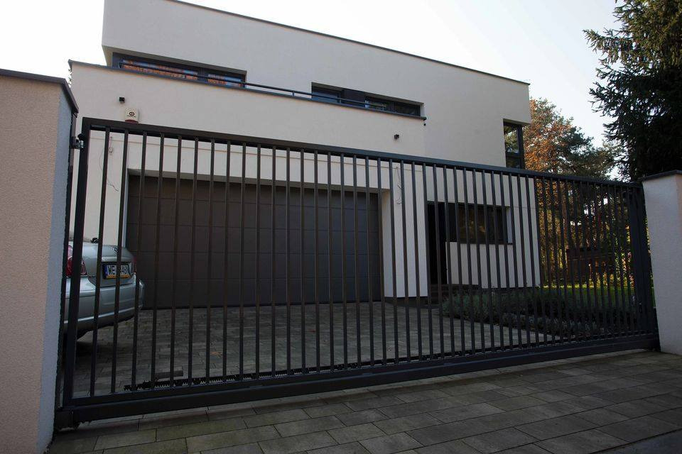 Nowoczesna posesja z przesuwną bramą wjazdową z metalowymi pionowymi prętami, widoczny fragment samochodu za bramą i brama garażowa w tle.