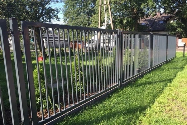 Nowoczesne ogrodzenie metalowe z pionowymi prętami i panelami, widok z zewnątrz, z zielonym trawnikiem i krzewami w tle.