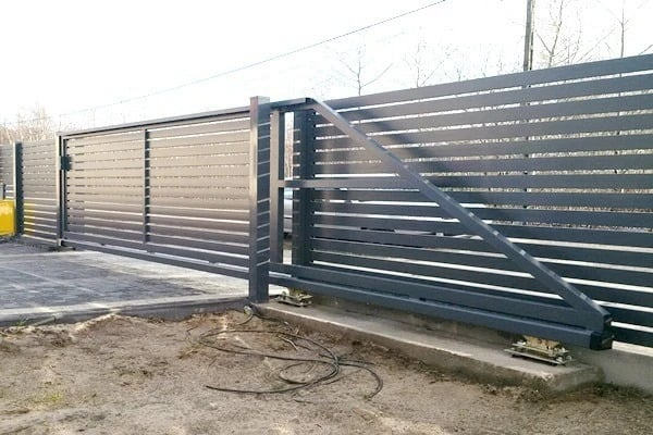 Szara, przesuwna brama wjazdowa z poziomymi panelami, widok z boku, z widocznymi elementami konstrukcyjnymi i betonową podstawą, na tle drzew bez liści.