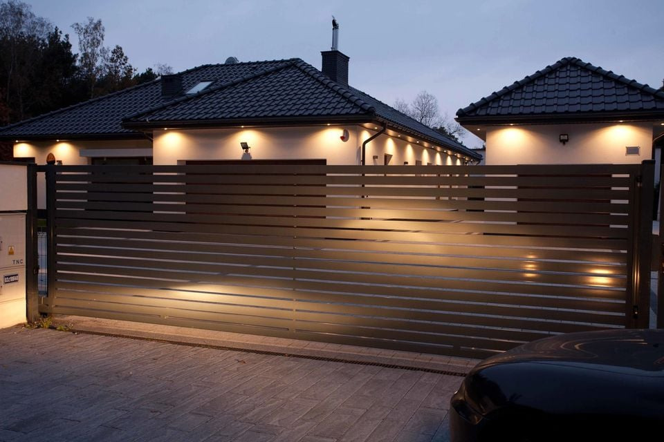 Automatyczna brama wjazdowa z poziomymi panelami, oświetlona reflektorami, na tle nowoczesnego domu z ciemną dachówką.