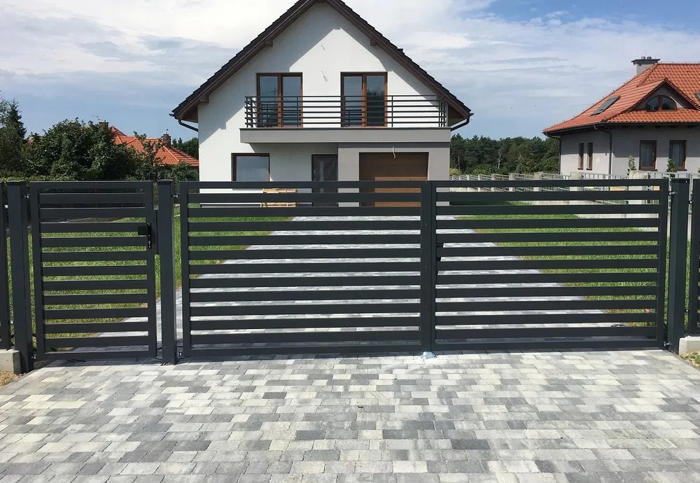 Nowoczesna, pozioma, metalowa brama wjazdowa i furtka w kolorze antracytowym na tle domu z balkonem i kostki brukowej.