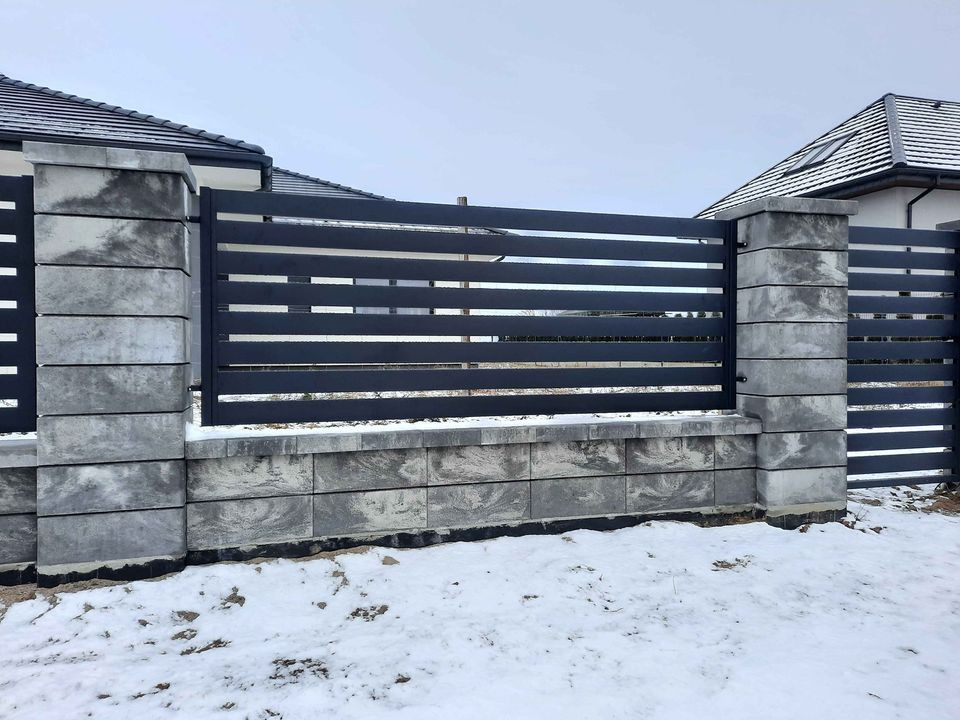 Nowoczesne, poziome przęsła ogrodzeniowe z ciemnego metalu, osadzone na słupkach z szarej kostki betonowej, częściowo pokryte śniegiem.