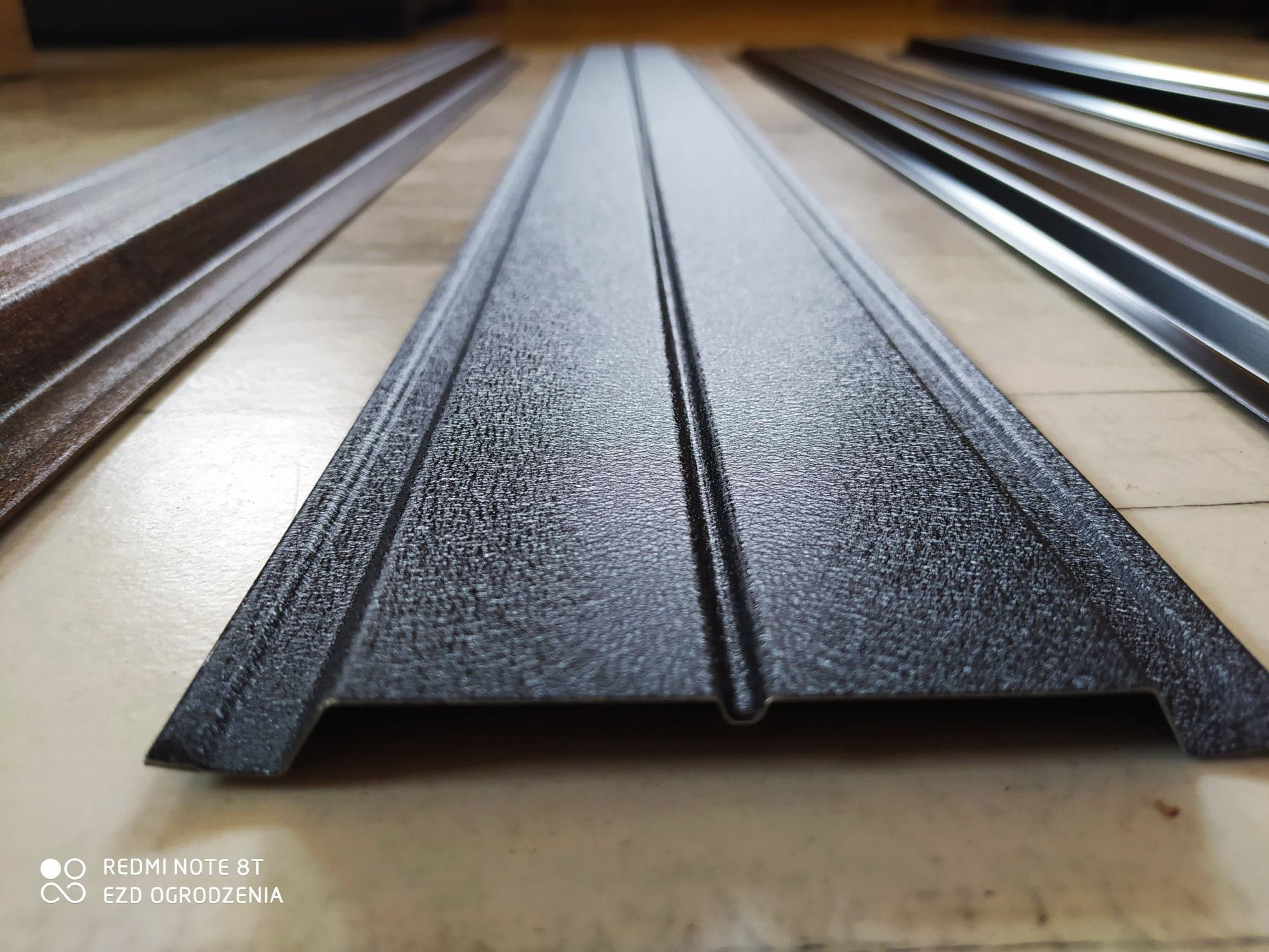 Profilowane metalowe panele ogrodzeniowe w kolorze grafitowym i brązowym, ułożone na podłodze, widoczne z bliska.