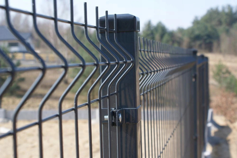 Fragment grafitowego ogrodzenia z paneli z widocznym słupkiem mocującym i zagięciami paneli, na tle rozmytej zieleni i zabudowań.