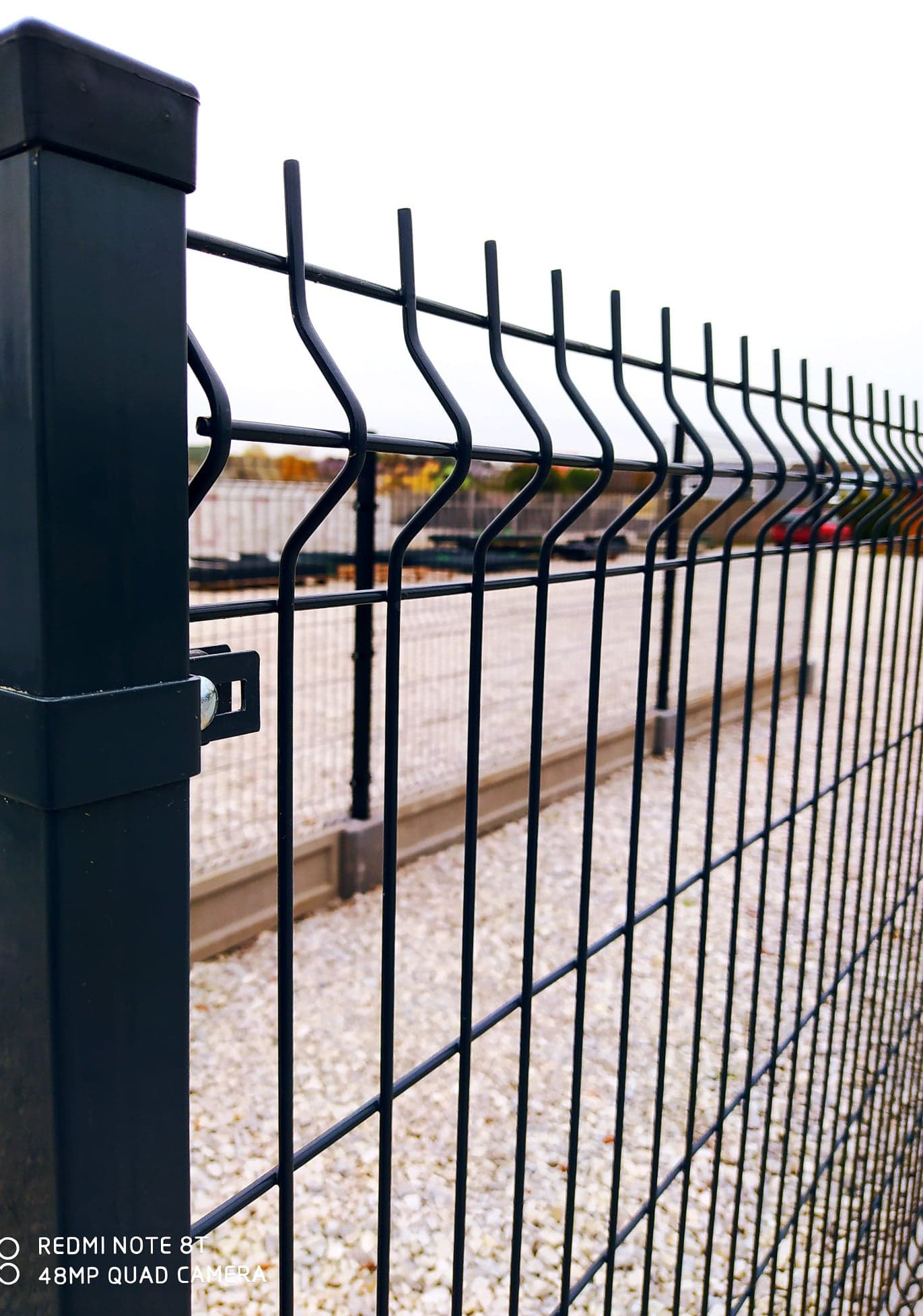 Ciemnoszare ogrodzenie panelowe z metalowymi słupkami na żwirowym podłożu, widok z bliska na mocowanie panela do słupka.