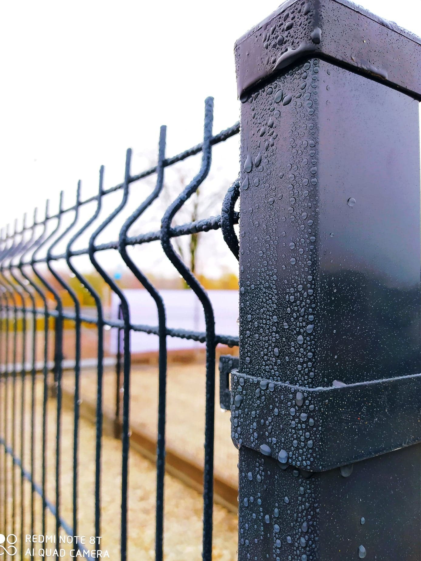 Czarny panel ogrodzeniowy z metalowymi prętami, pokryty kroplami wody, widoczny słupek ogrodzeniowy z nasadką, w tle rozmyty krajobraz.