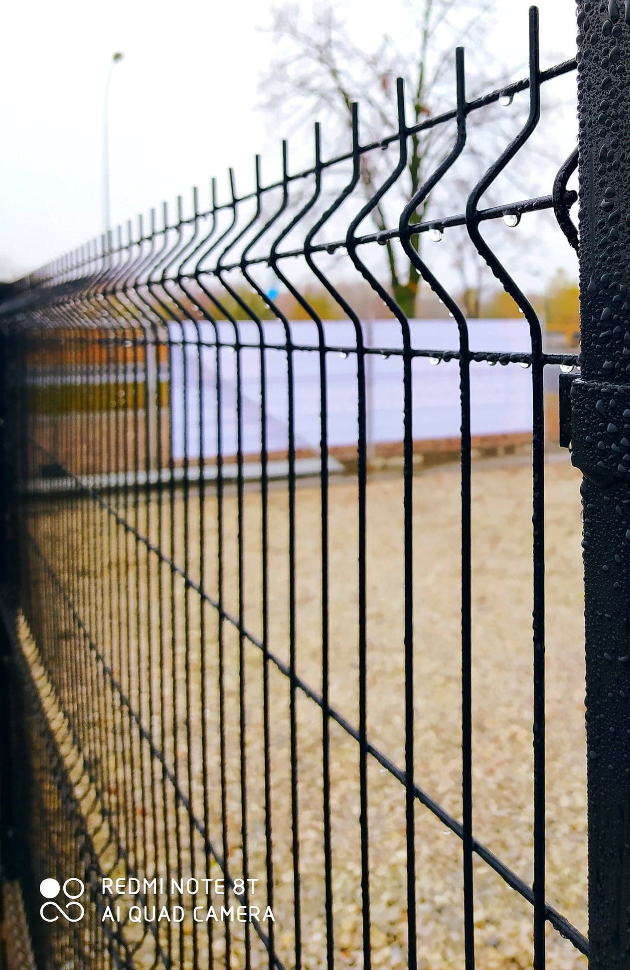 Czarne, metalowe ogrodzenie panelowe w Zduńskiej Woli, pokryte kroplami deszczu, z widocznym w tle kortem tenisowym i drzewem bez liści.