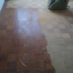 Raf -Tob remontujemy - Częściowo wyszlifowany parkiet w jodełkę, z widoczną szlifierką pod oknem. Po lewej stronie parkiet w ciemnym odcieniu, po prawej jaśniejszy, surowy odcień drewna.