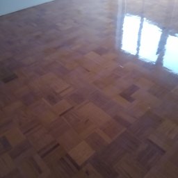 Raf -Tob remontujemy - Błyszcząca, lakierowana podłoga parkietowa w jodełkę, odbijająca światło z okna. Widoczne detale ułożenia klepek i refleksy świetlne. Świeżo wykończona powierzchnia.