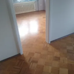 Parkiet ułożony w kwadraty, widoczny z perspektywy przejścia między pokojami. W tle biały grzejnik i ściana w jasnym odcieniu fioletu.