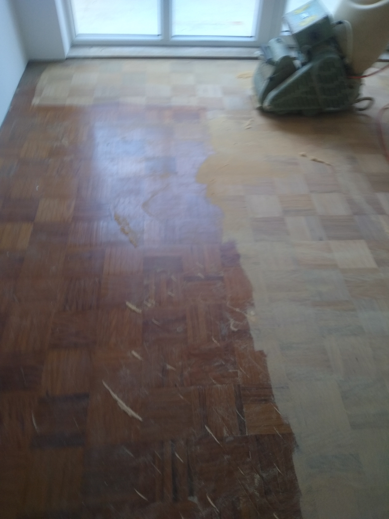 Częściowo wyszlifowany parkiet w jodełkę, z widoczną szlifierką pod oknem. Po lewej stronie parkiet w ciemnym odcieniu, po prawej jaśniejszy, surowy odcień drewna.