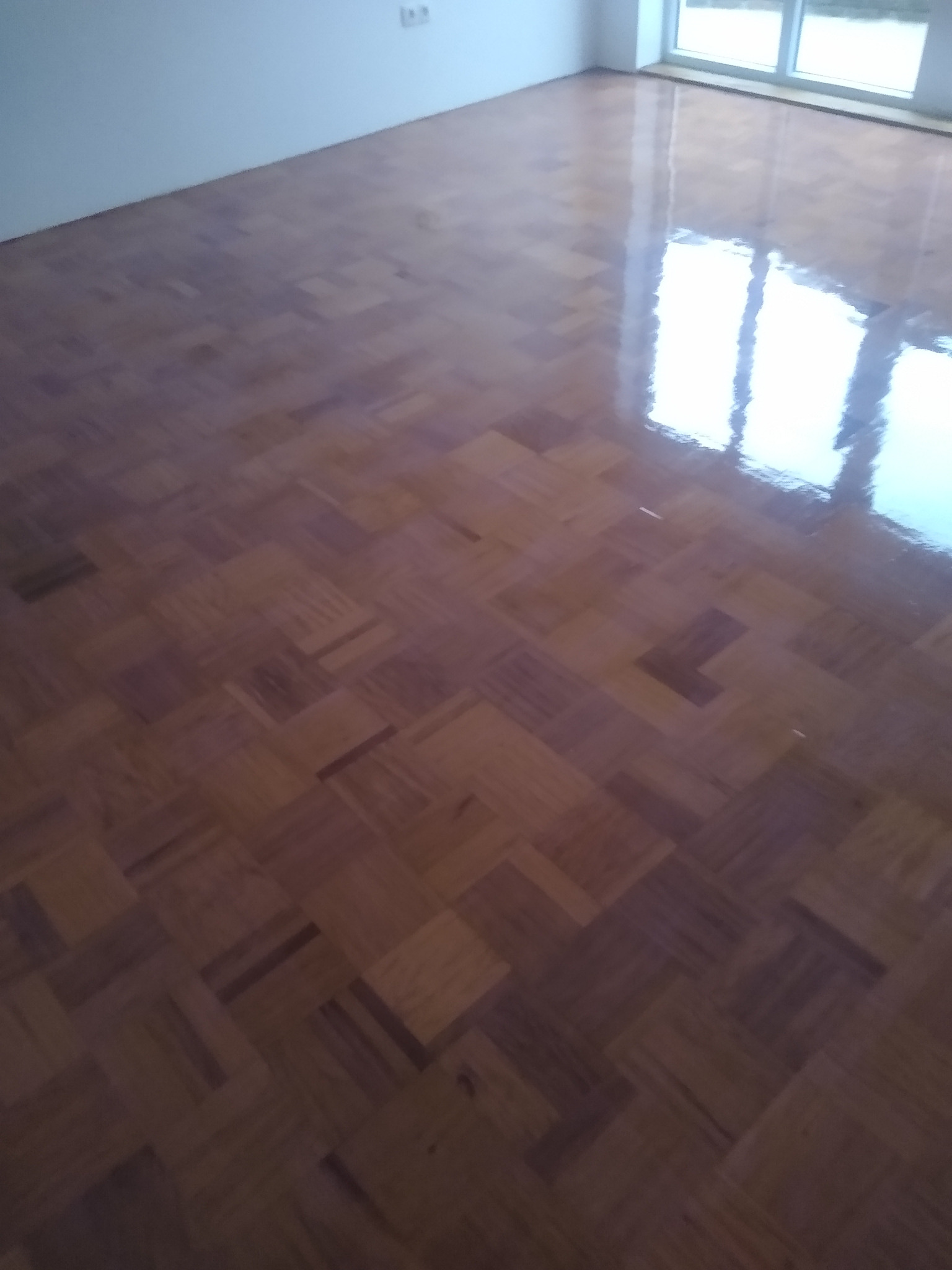 Błyszcząca, lakierowana podłoga parkietowa w jodełkę, odbijająca światło z okna. Widoczne detale ułożenia klepek i refleksy świetlne. Świeżo wykończona powierzchnia.