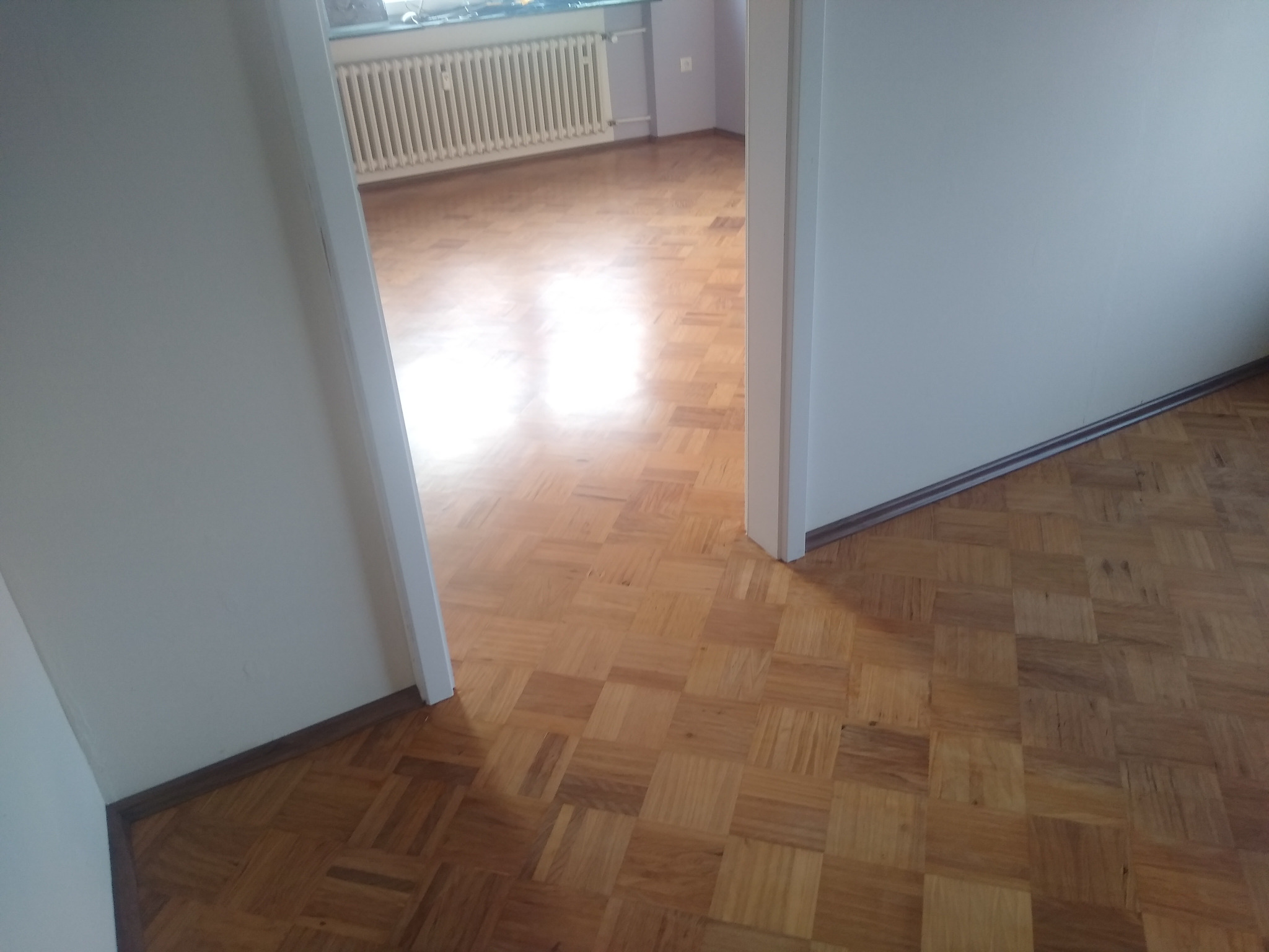 Parkiet ułożony w kwadraty, widoczny z perspektywy przejścia między pokojami. W tle biały grzejnik i ściana w jasnym odcieniu fioletu.