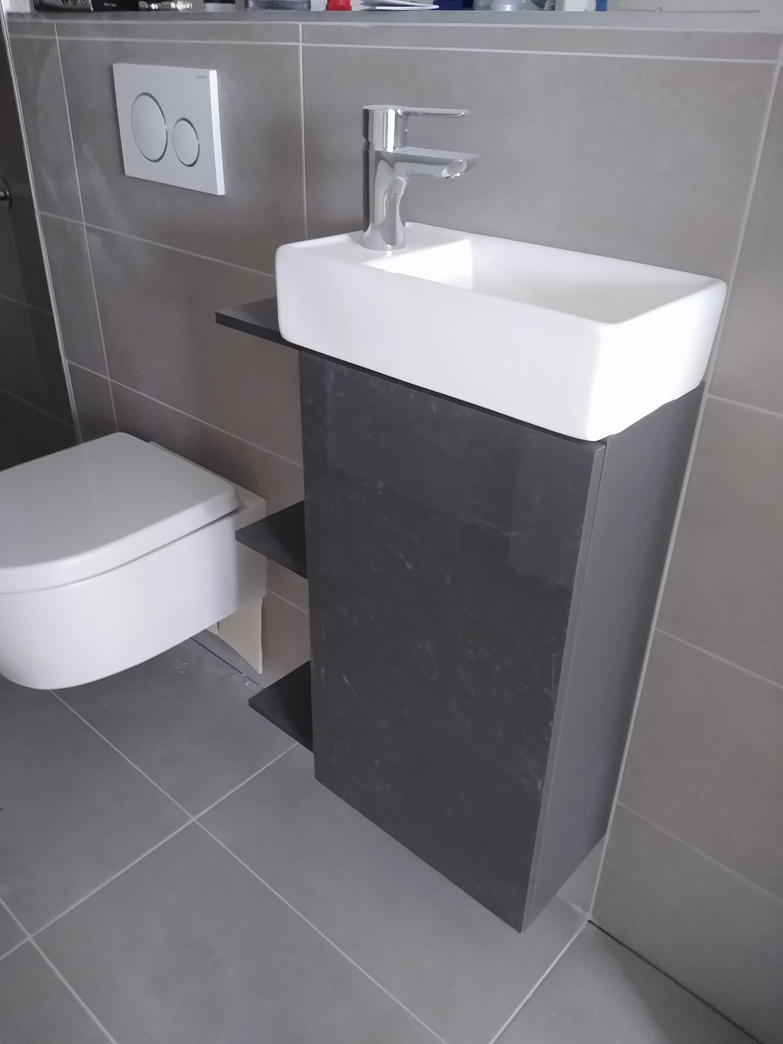 Nowoczesna, szara łazienka z podwieszaną toaletą i umywalką na szafce z półkami. Minimalistyczny design, szare płytki, chromowana bateria.