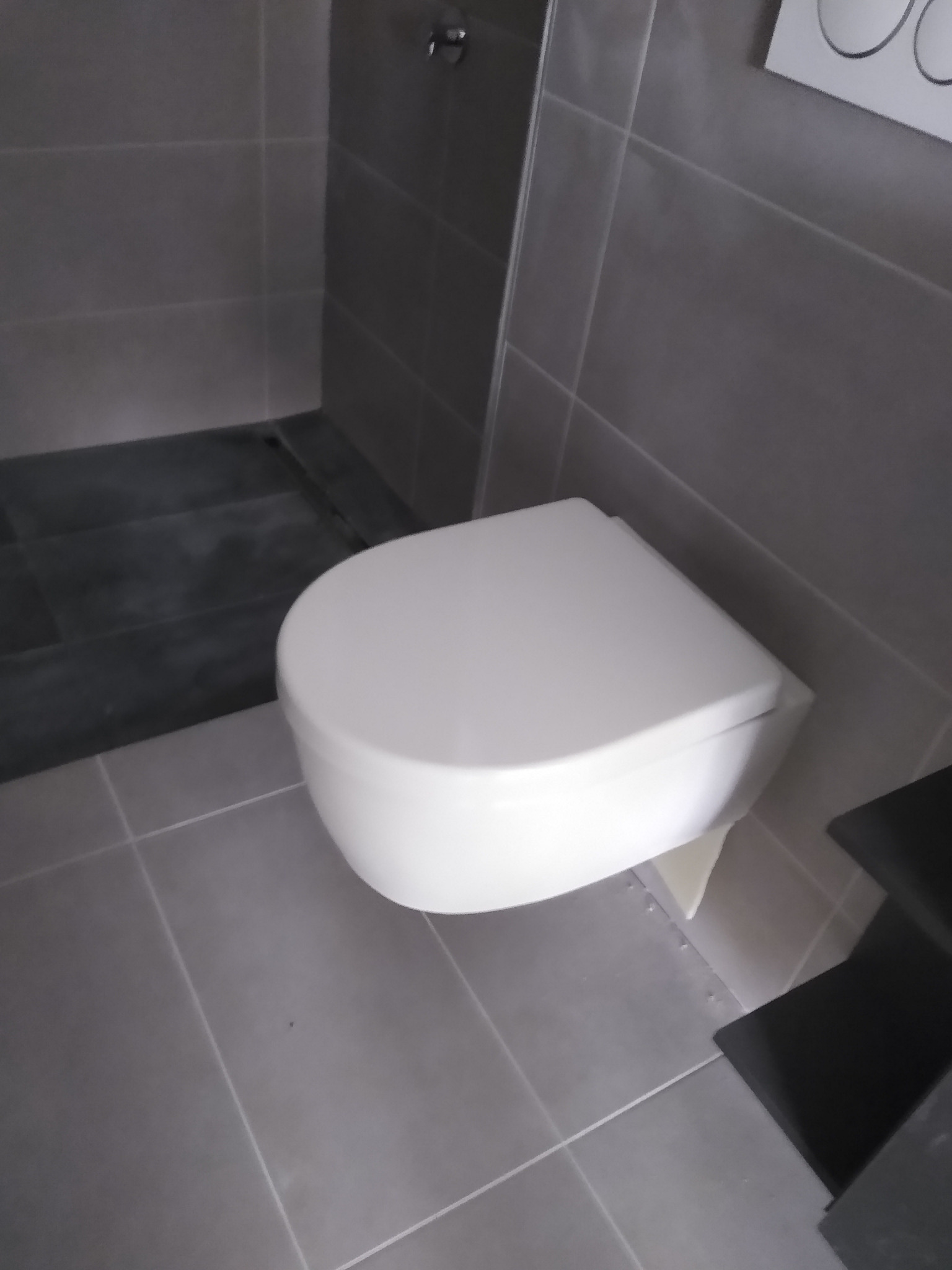 Nowoczesna, podwieszana toaleta w łazience wykończonej szarymi płytkami. Widoczne elementy armatury i fragment półki.