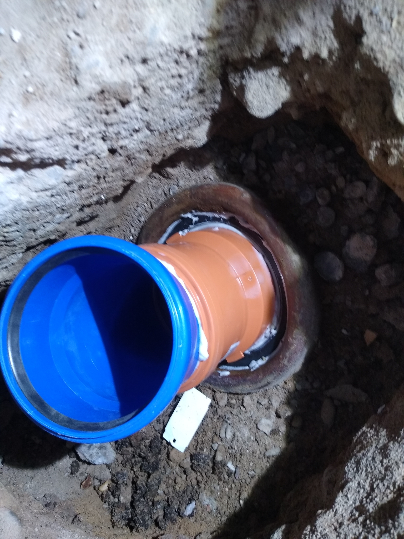 Szczegółowe ujęcie pomarańczowej rury kanalizacyjnej z niebieskim zakończeniem, osadzonej w ziemi, ukazujące etap prac instalacyjnych z użyciem uszczelniacza.