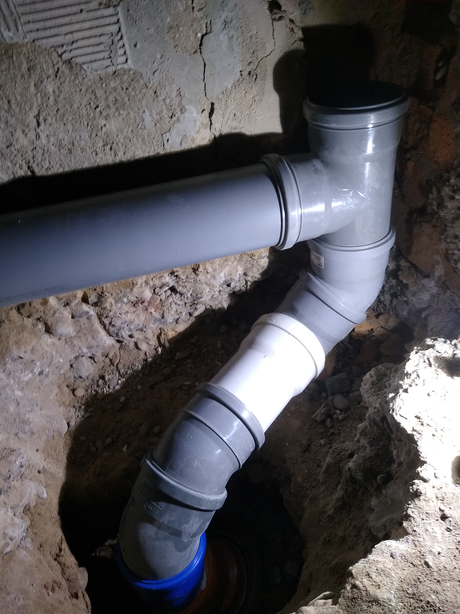 Szare rury kanalizacyjne ułożone w wykopie ziemnym, z widocznymi elementami instalacji i fragmentem starej ściany z cegieł w tle.