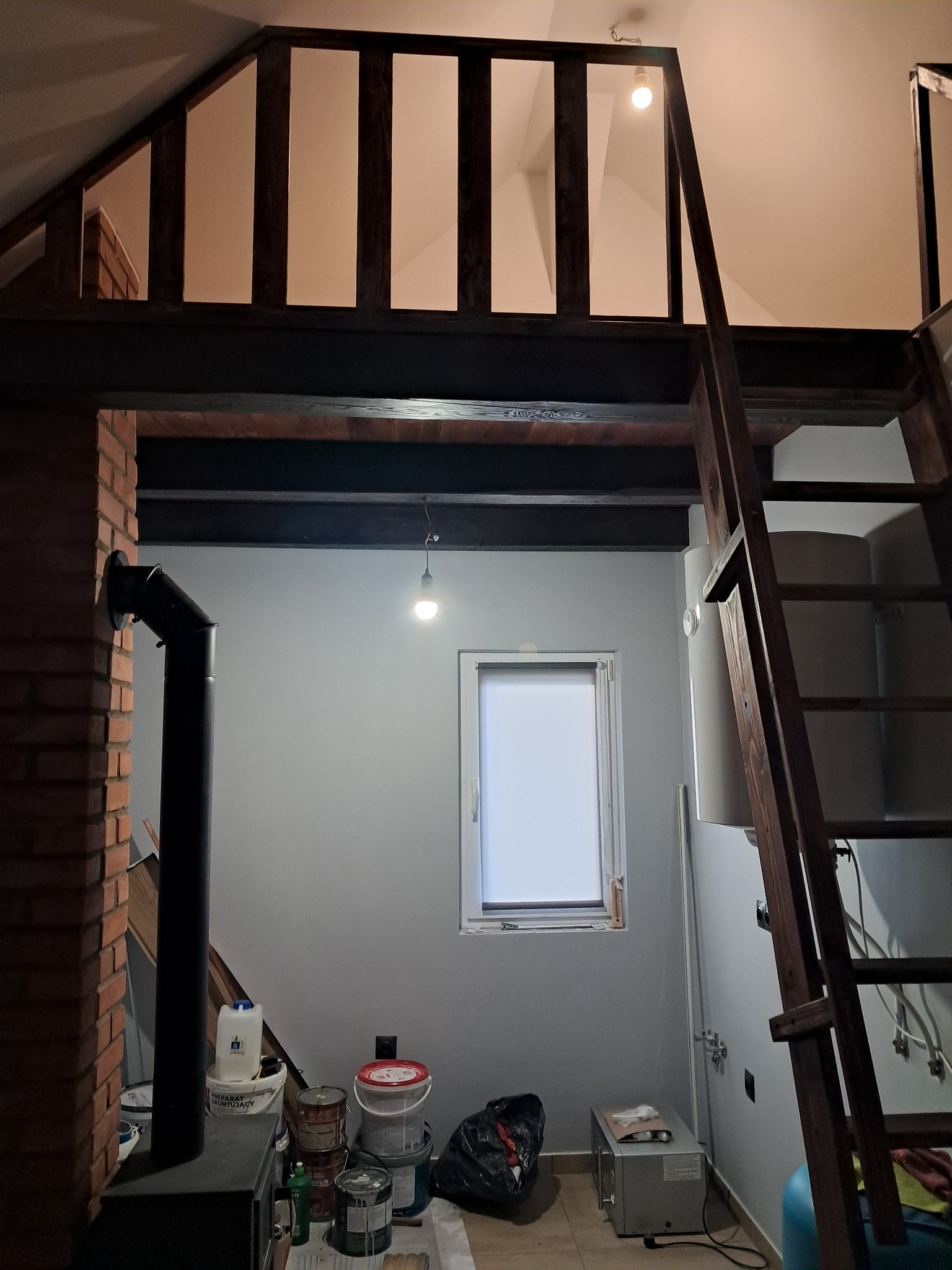 Wnętrze pokoju z antresolą w trakcie prac wykończeniowych. Widoczne farby, drabina, piec i okno z roletą. Surowy stan, przygotowanie do dalszych prac.