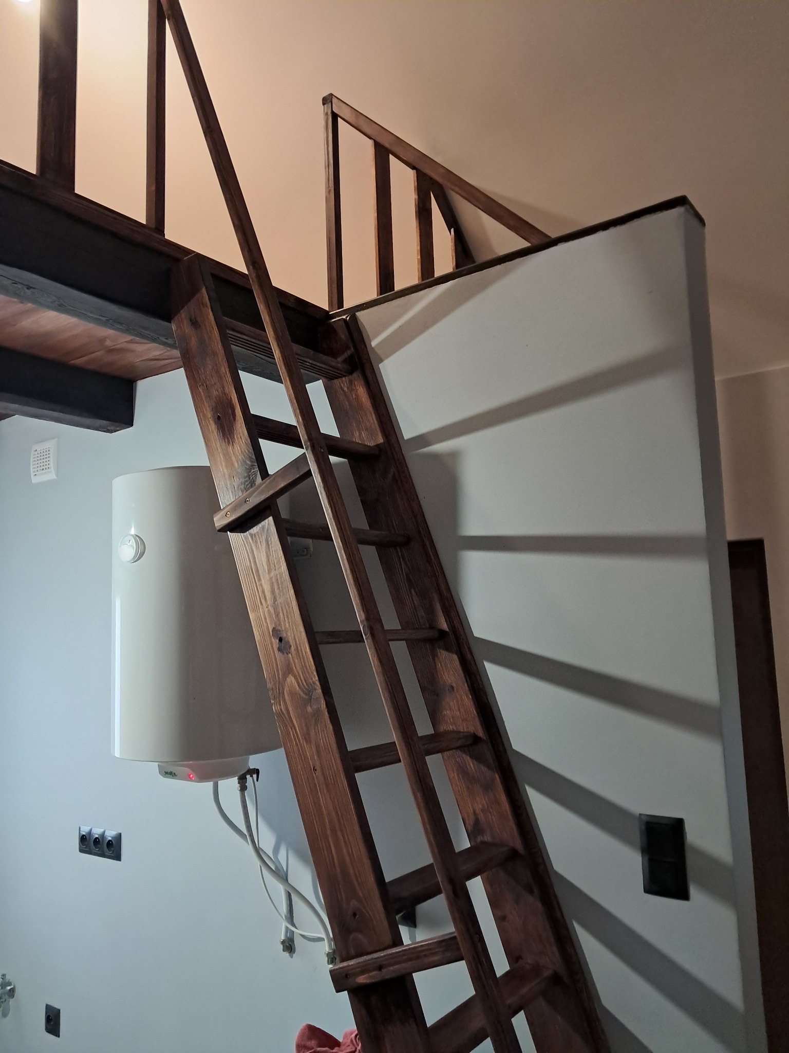 Drewniana drabina prowadząca na antresolę w mieszkaniu w Brzozówce. Widoczny bojler i gniazdka elektryczne na białej ścianie. Ujęcie z dołu, podkreślające wysokość.