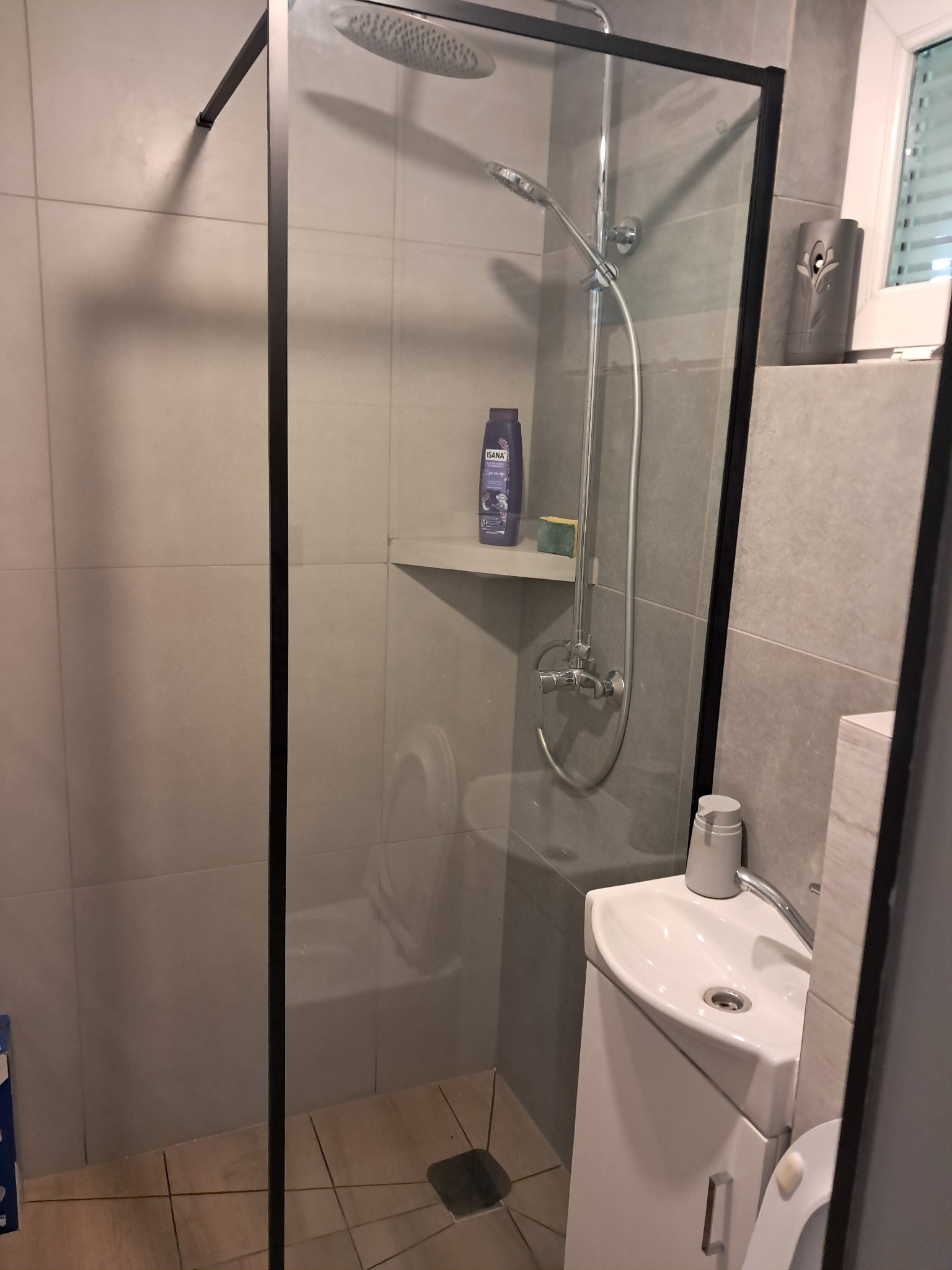 Nowoczesna kabina prysznicowa z czarną ramą i szarymi płytkami. Widoczna umywalka narożna i półka z kosmetykami. Minimalistyczny design łazienki.