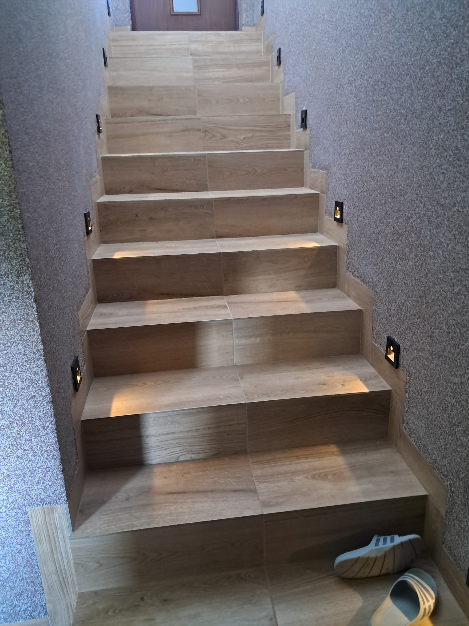 Wykończone schody z drewnopodobnych płytek z oświetleniem punktowym w ścianie o fakturze baranka. Na dole leżą klapki.