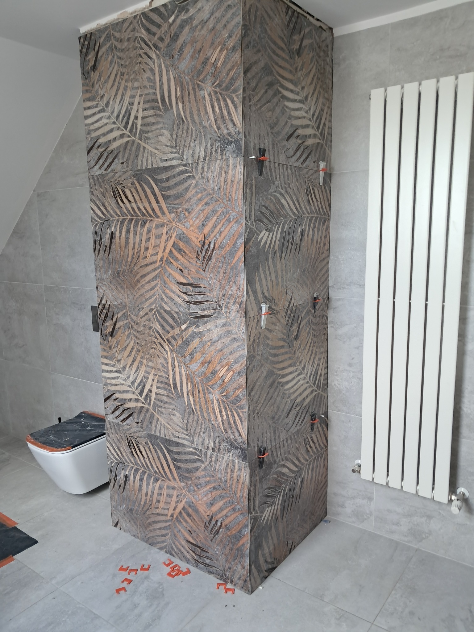 Wykończona obudowa WC z płytek w liście palmy, z krzyżykami dystansowymi, w szarej łazience z grzejnikiem żeberkowym. Nowoczesny design z motywem roślinnym.