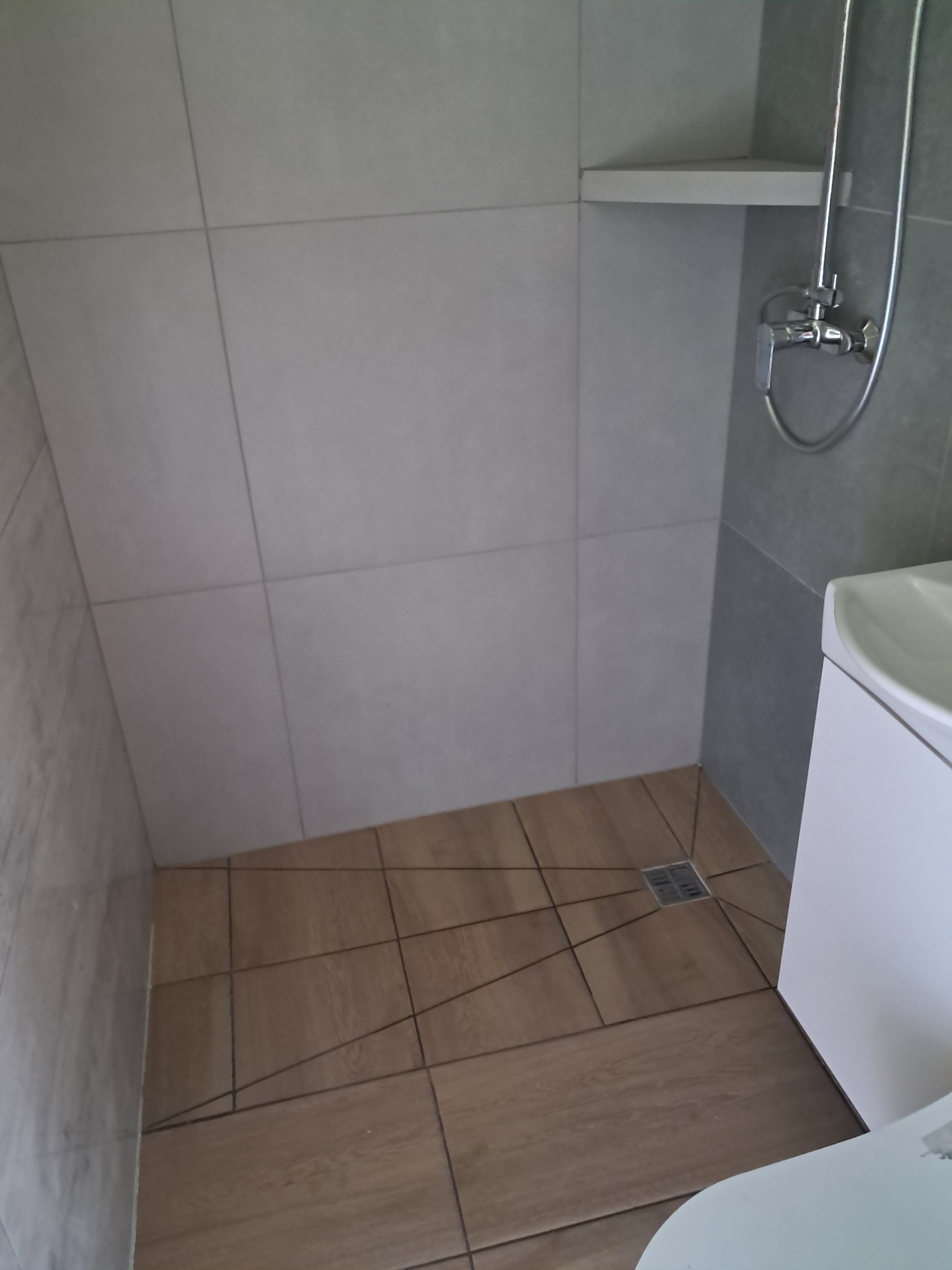 Nowoczesna kabina prysznicowa z drewnopodobną podłogą i szarymi płytkami ściennymi. Minimalistyczny design z odpływem liniowym i półką.