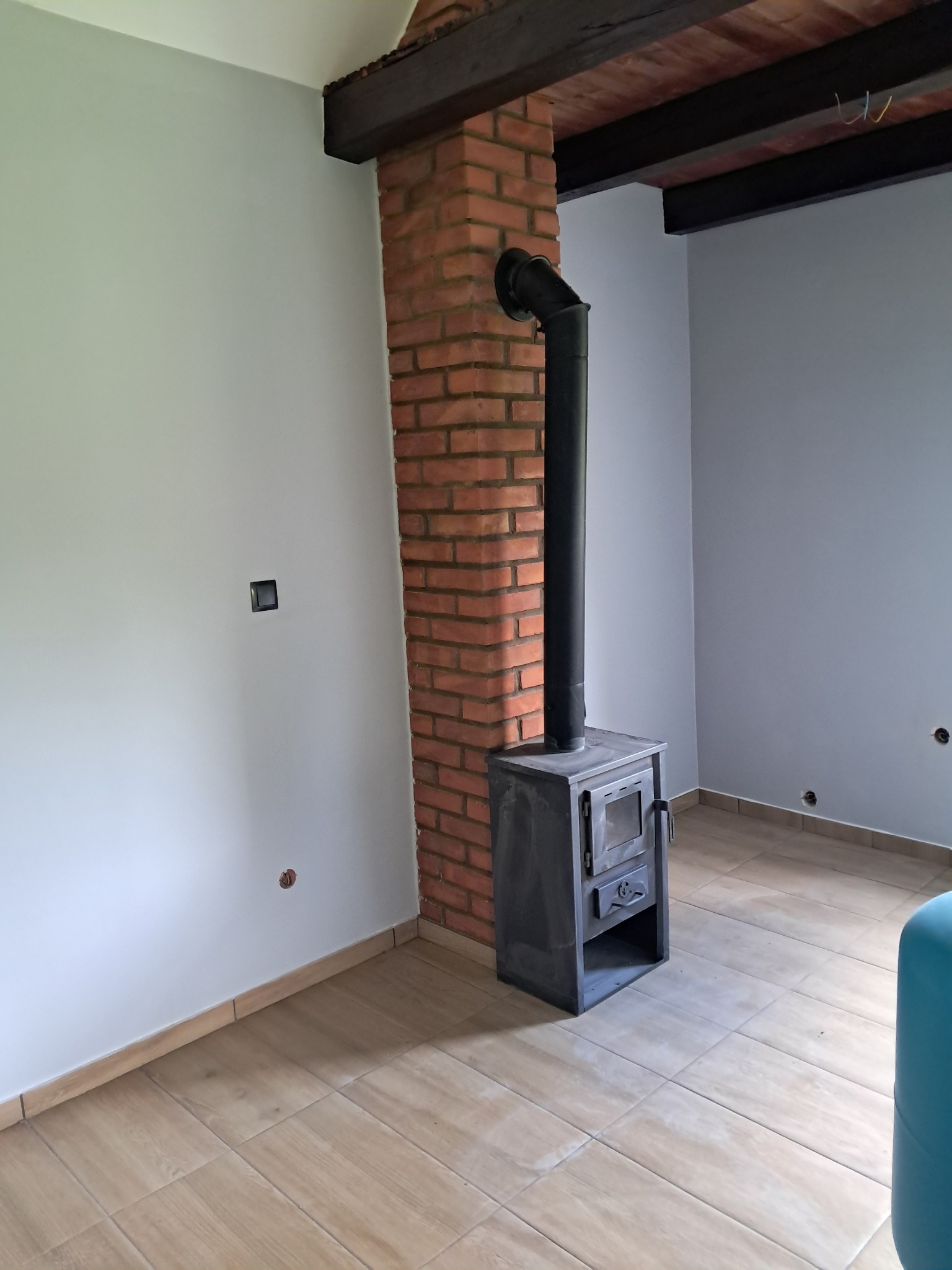 Wnętrze z piecem wolnostojącym i ceglanym kominem. Jasne ściany, drewniane belki stropowe i podłoga z płytek imitujących drewno. Widoczne gniazdka elektryczne.