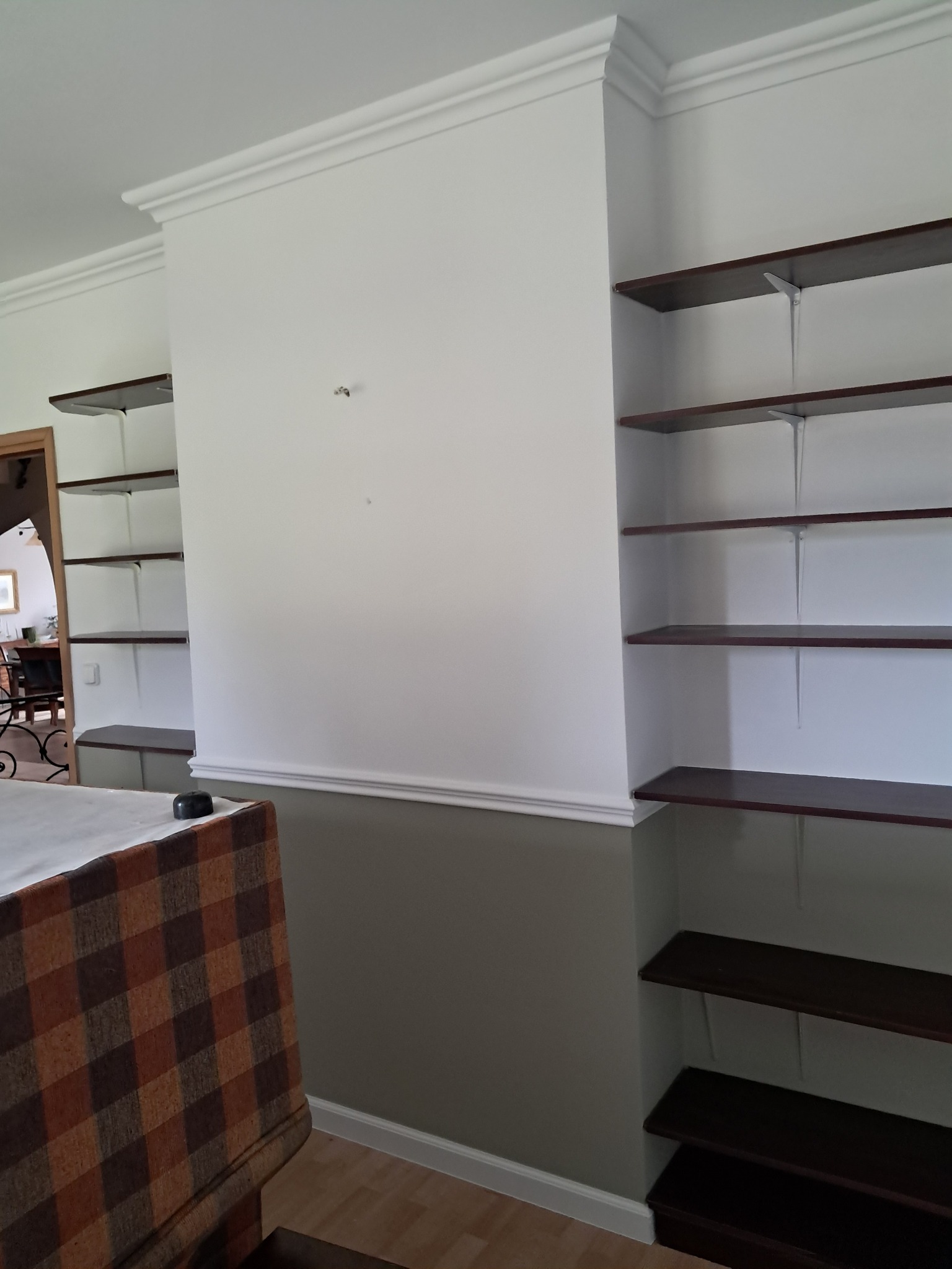 Biała ściana z dekoracyjną listwą i wbudowanymi, ciemnymi półkami. Dolna część ściany pomalowana na szaro. Widoczny fragment mebla z wzorzystą tapicerką.