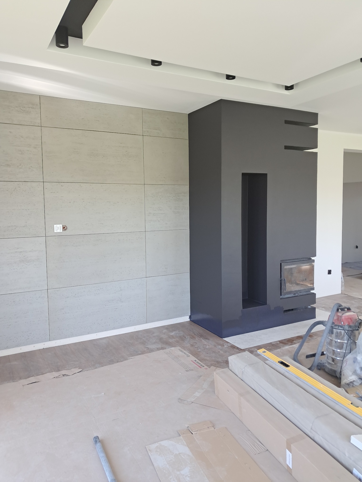 Wnętrze w trakcie wykańczania: szara ściana z płytek, kominek w zabudowie, podłoga zabezpieczona kartonem, widoczne narzędzia i materiały budowlane. Oświetlenie sufitowe.