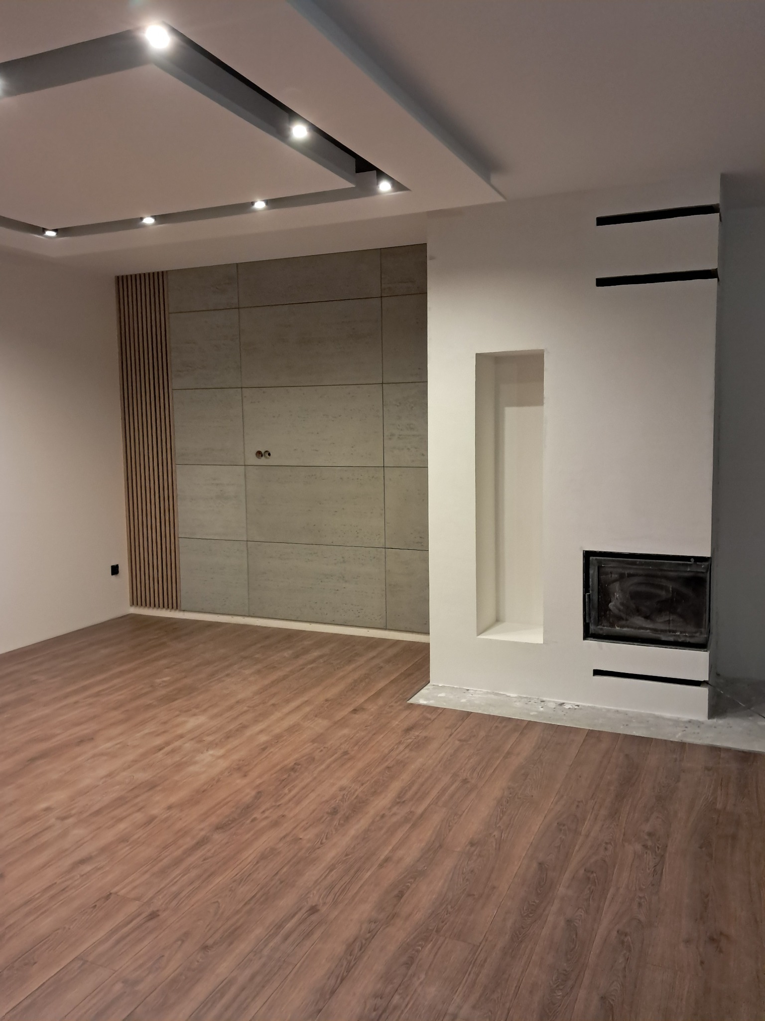 Wykończone wnętrze pokoju z drewnianą podłogą, ścianą z paneli i płyt oraz kominkiem z wnęką. Sufit podwieszany z oświetleniem punktowym.