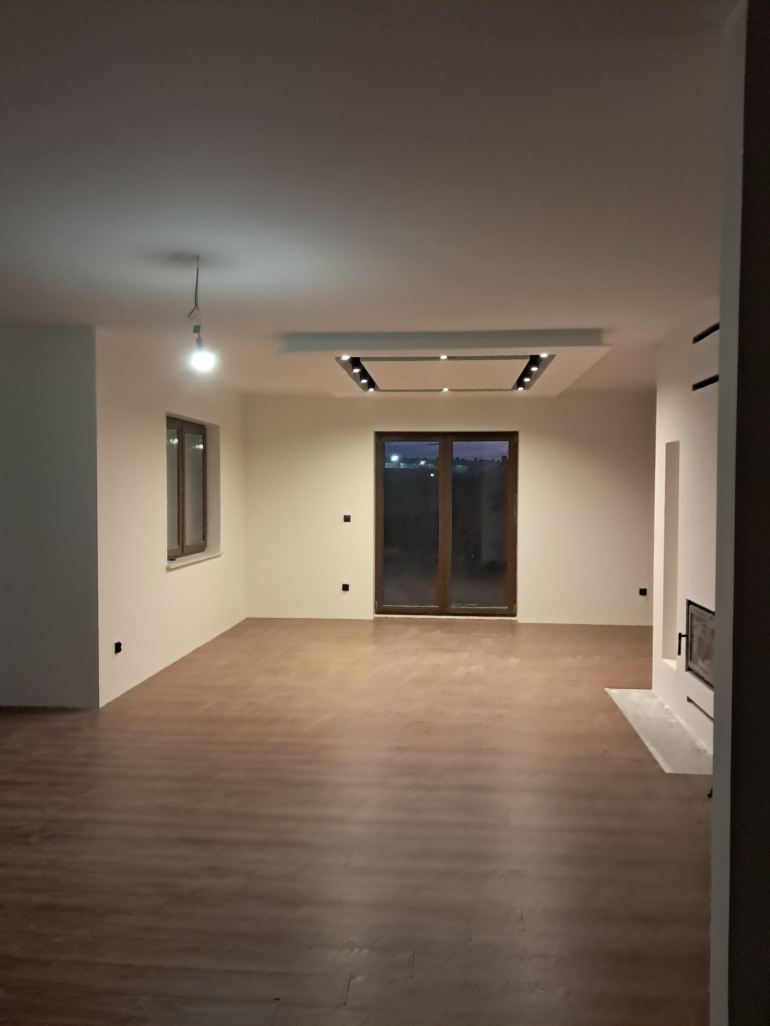 Jasne wnętrze z panelami podłogowymi, oświetleniem sufitowym i kominkiem. Widoczne okno i drzwi balkonowe. Minimalistyczny design.