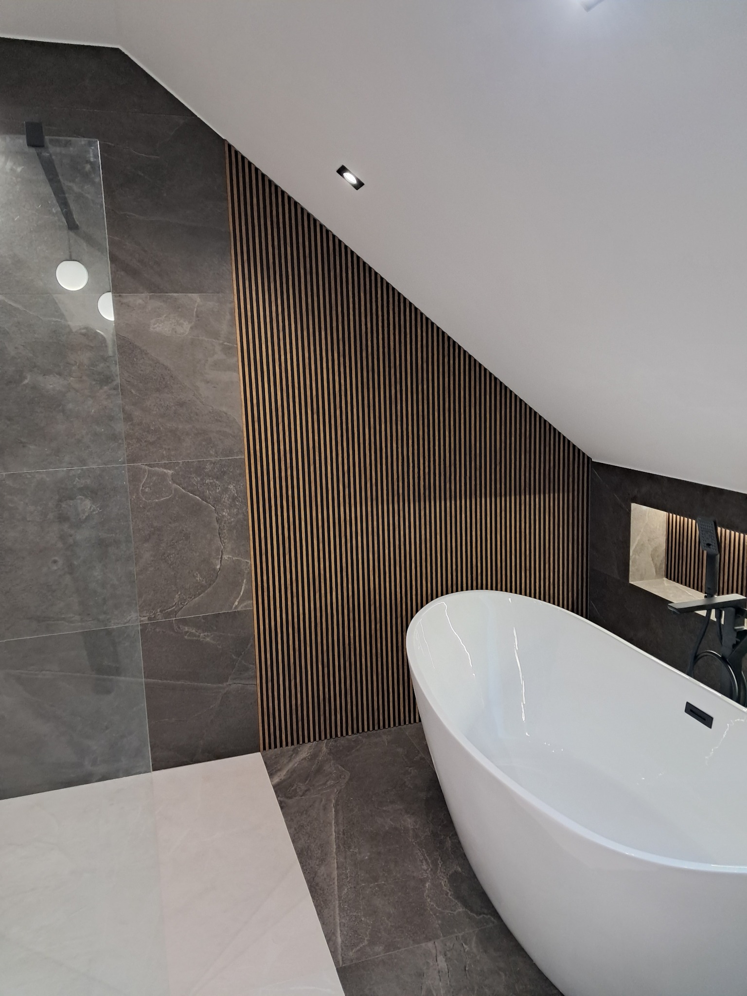 Nowoczesna łazienka na poddaszu z wolnostojącą wanną, szarą glazurą imitującą marmur i drewnianymi lamelami na skośnej ścianie. Minimalistyczne oświetlenie punktowe.