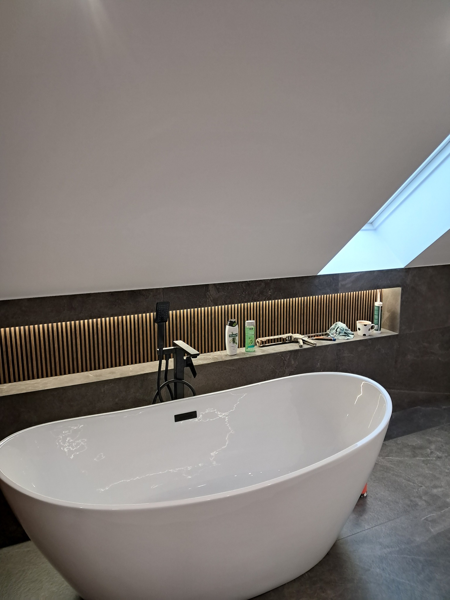 Nowoczesna łazienka na poddaszu z wolnostojącą białą wanną, czarną armaturą i półką z oświetleniem LED. Widoczne narzędzia i kosmetyki na półce.