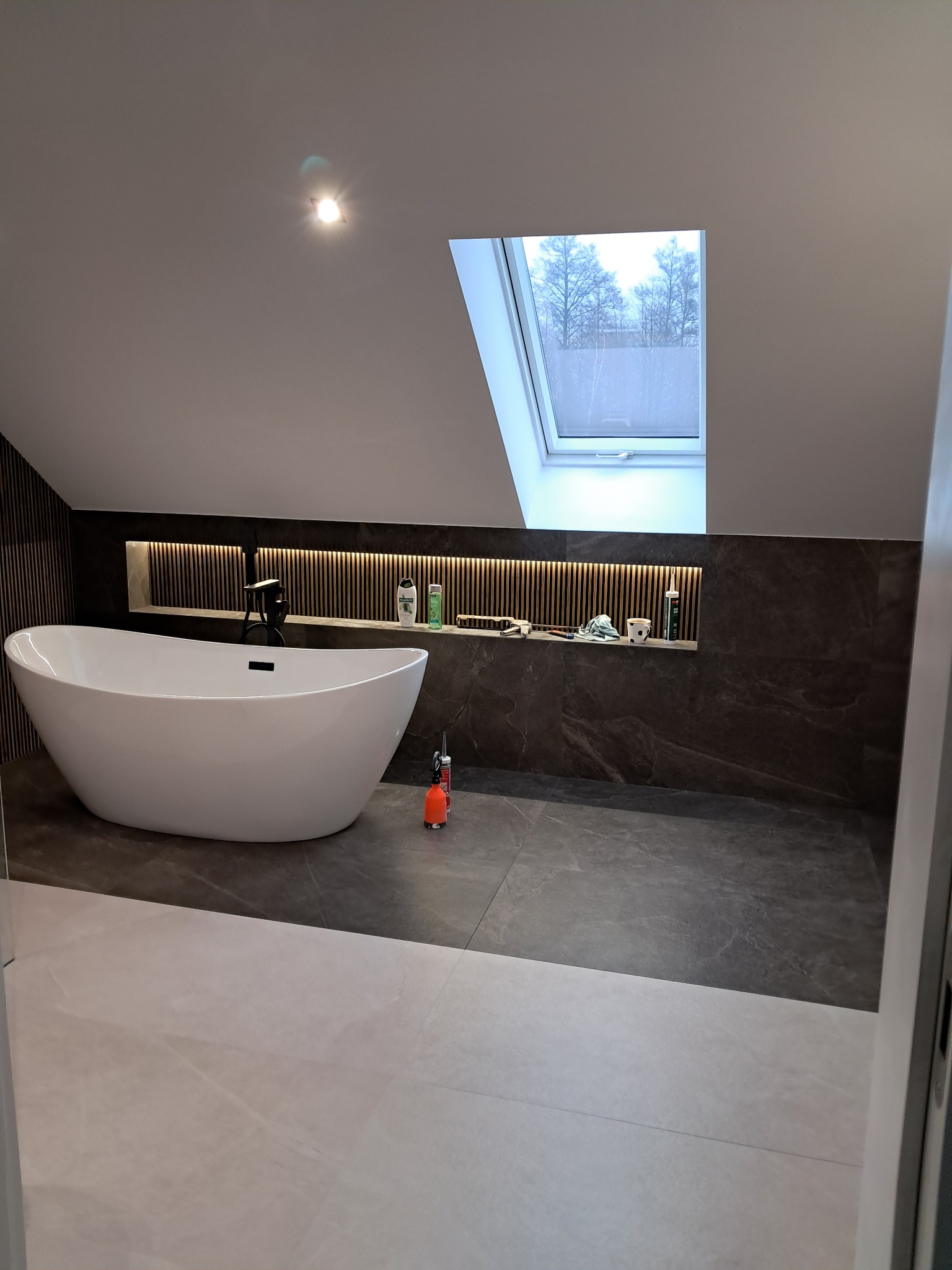 Nowoczesna łazienka na poddaszu z wolnostojącą wanną, szarą glazurą i oknem dachowym. Wnęka z oświetleniem LED i dekoracyjnymi elementami. Minimalistyczny design.