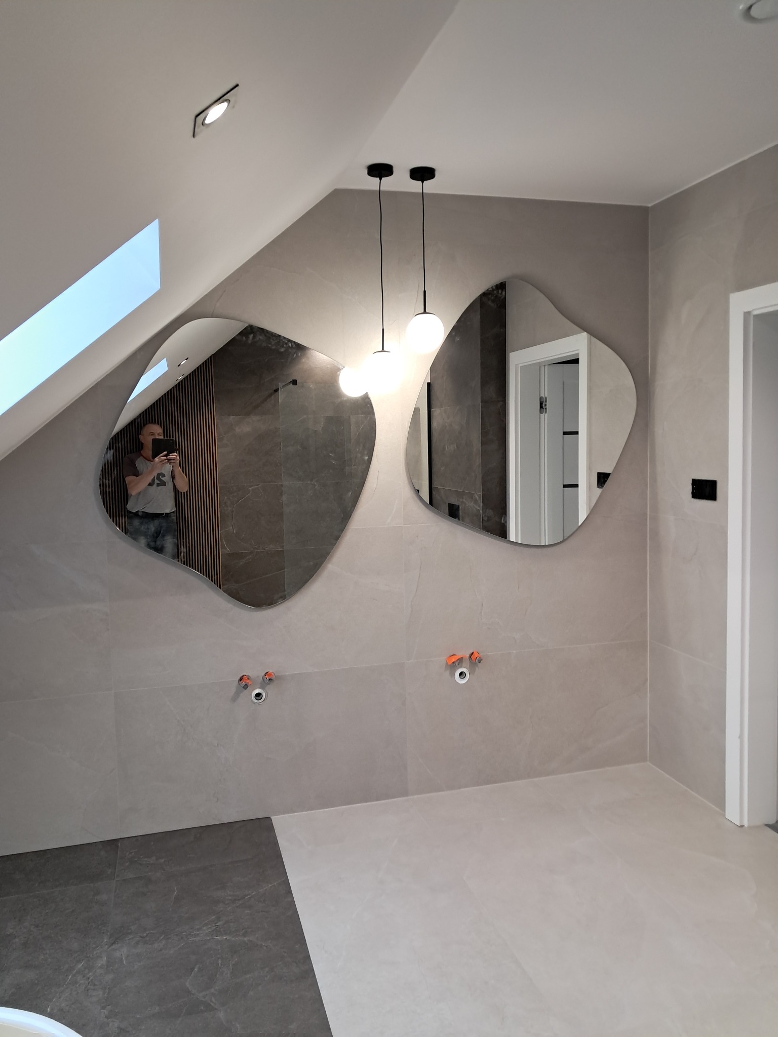 Wykończona łazienka na poddaszu z dwoma asymetrycznymi lustrami i szarą glazurą. Widoczne przyłącza wodne i nowoczesne oświetlenie sufitowe.