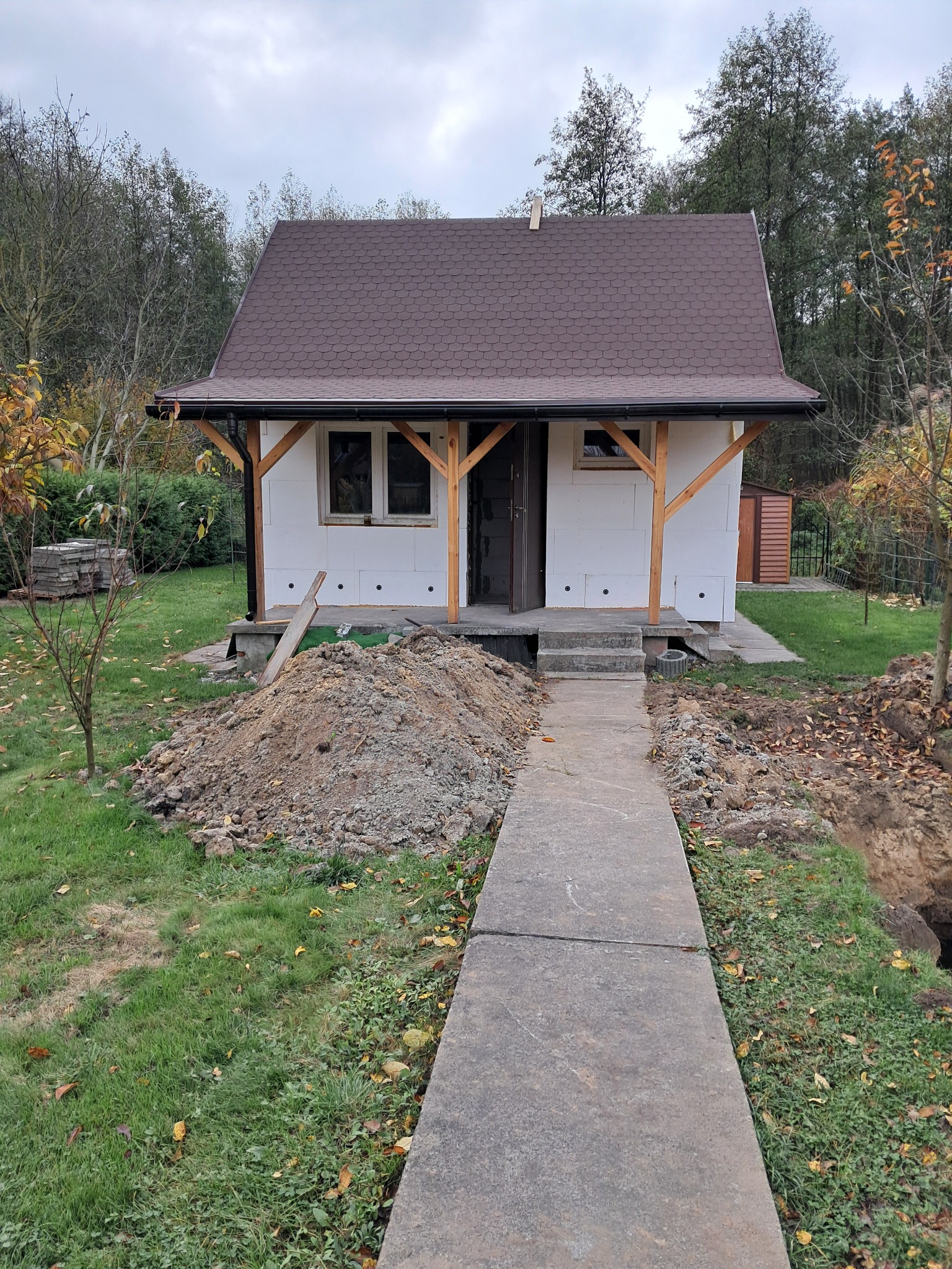 Niedokończony budynek z brązowym dachem i białymi ścianami, widoczna sterta ziemi i betonowa ścieżka prowadząca do wejścia. Prace wykończeniowe w toku.