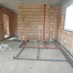 Surowy stan wnętrza budynku w trakcie rozprowadzania instalacji elektrycznej i rur, widoczne kable ułożone na podłodze i wyprowadzenia ze ścian z cegły.