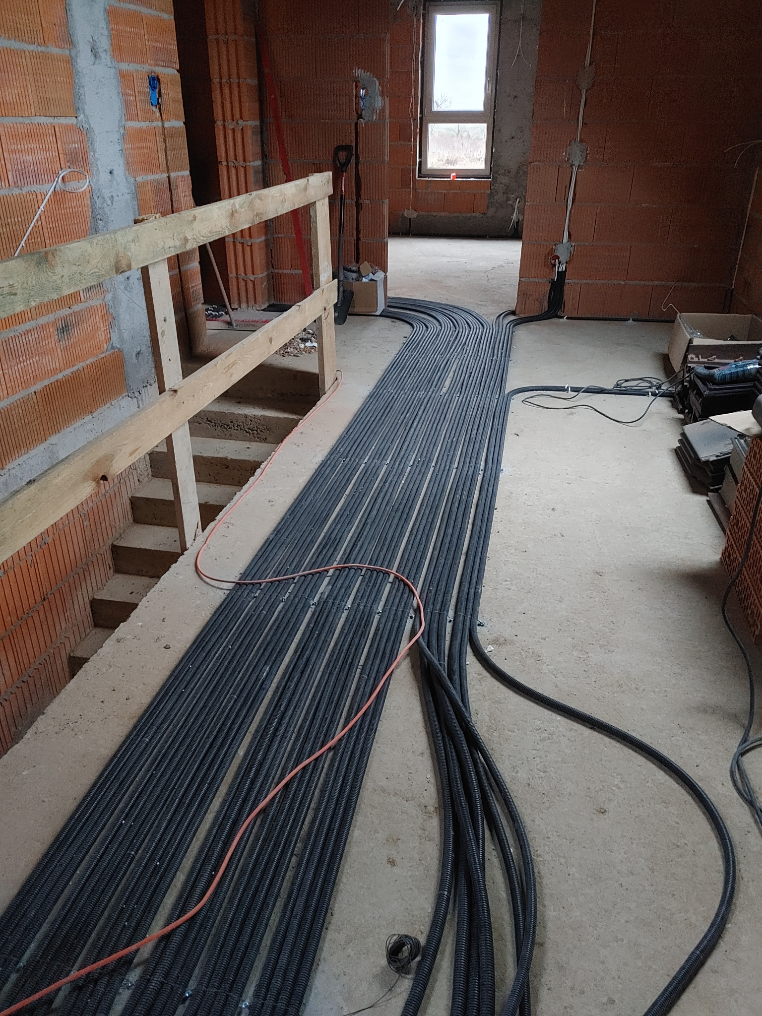 Równolegle ułożone rury karbowane instalacji elektrycznej na surowym podłożu w trakcie budowy, z widocznym oknem i ceglanymi ścianami.