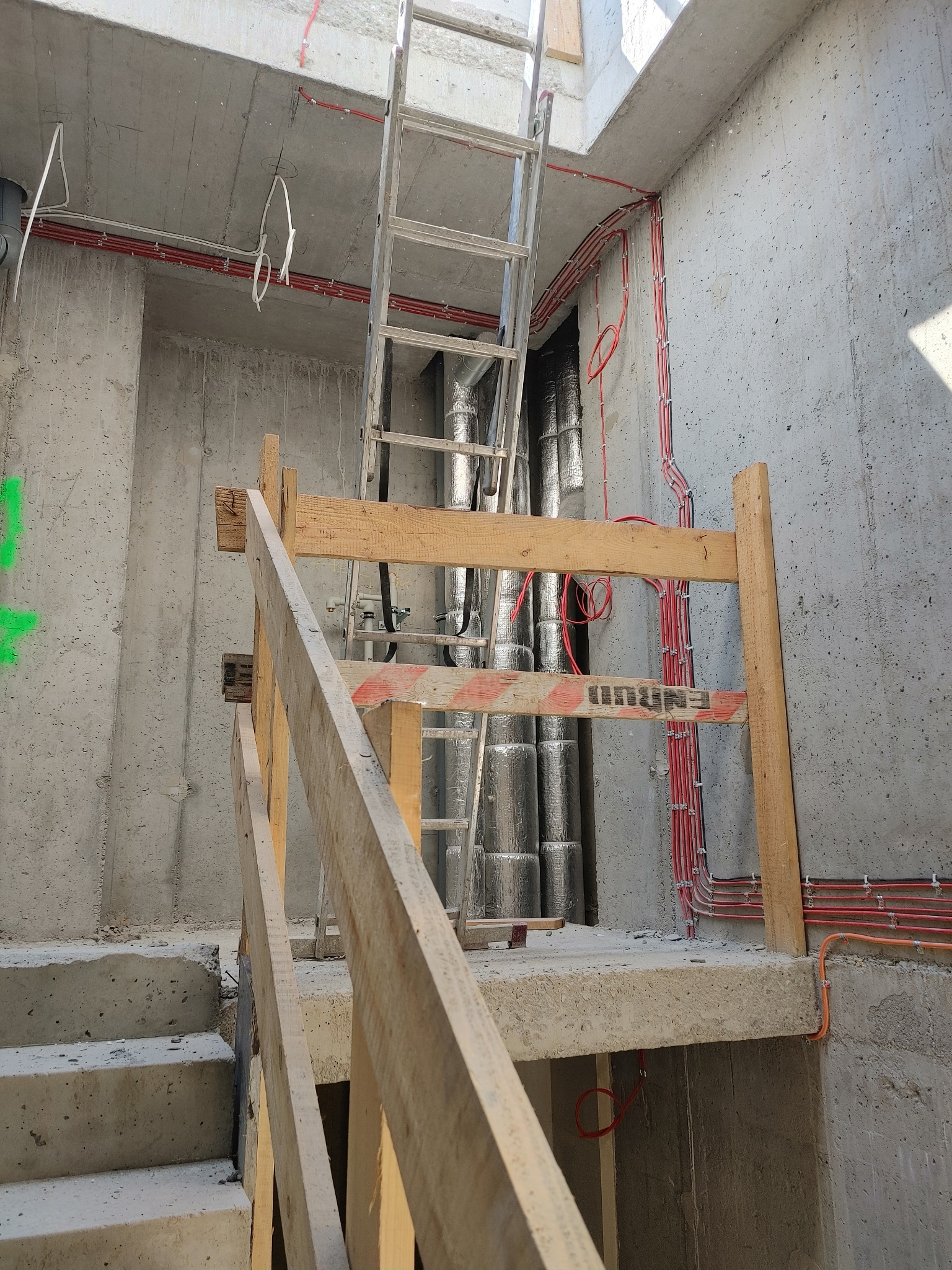 Surowy stan budynku w trakcie prac: widoczne kable elektryczne prowadzone po ścianie, pionowe rury w izolacji, drabina oparta o ścianę oraz prowizoryczne drewniane barierki zabezpieczające.