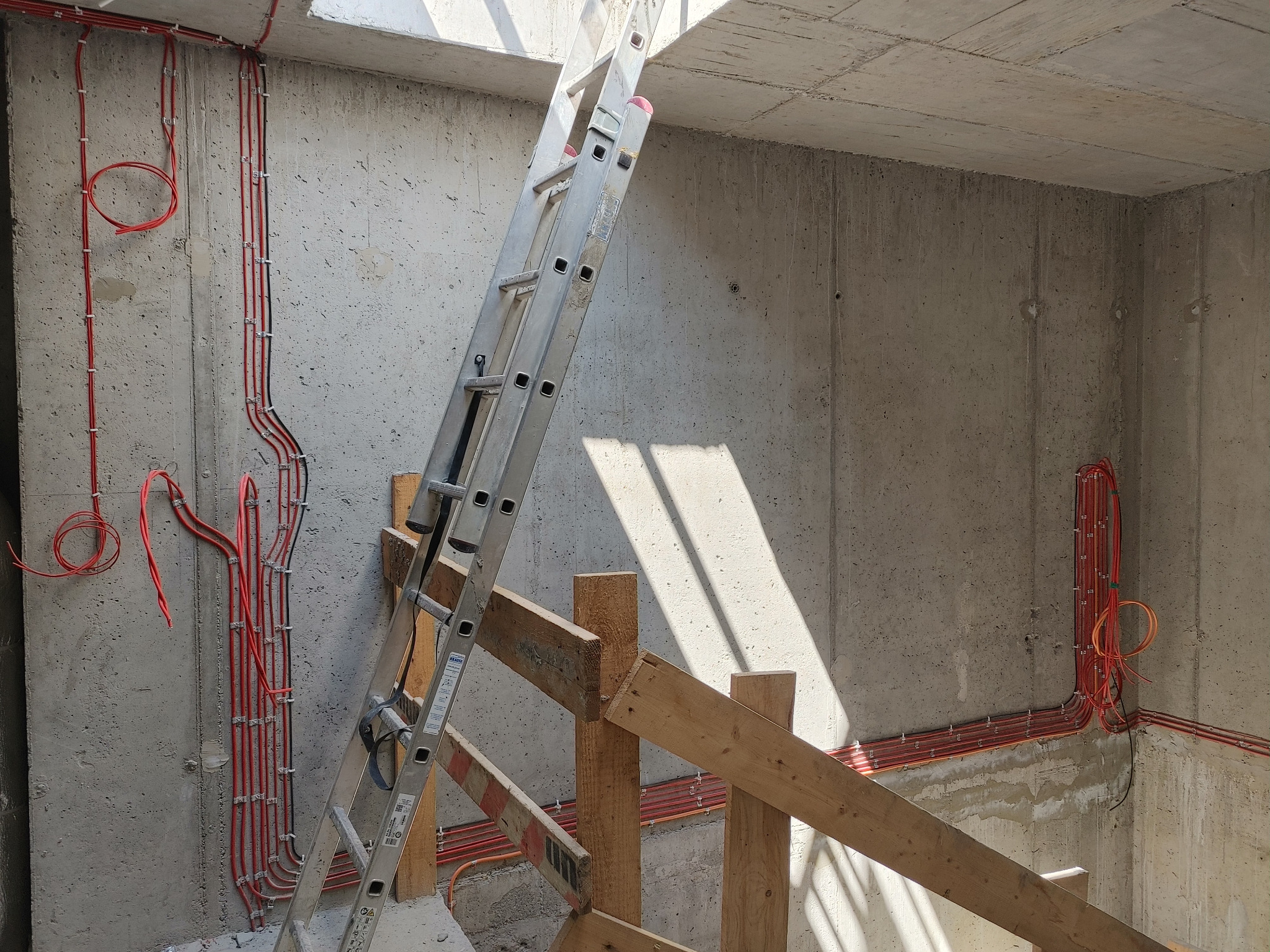 Surowy betonowy pokój w trakcie prac wykończeniowych, widoczne poprowadzone czerwone kable elektryczne, aluminiowa drabina oparta o drewniane rusztowanie, wpadające przez otwór w suficie światło.