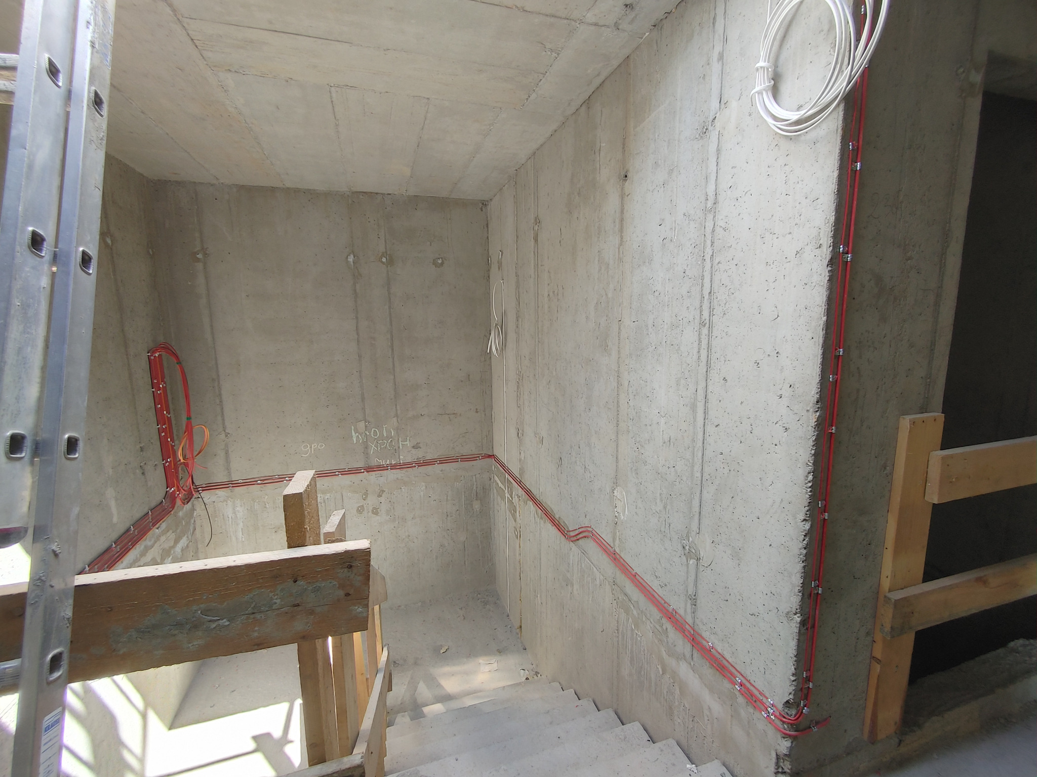Surowy betonowy korytarz schodowy w trakcie budowy z widocznymi czerwonymi kablami elektrycznymi przymocowanymi do ścian i białymi kablami zwisającymi z sufitu, zabezpieczony drewnianą barierką...