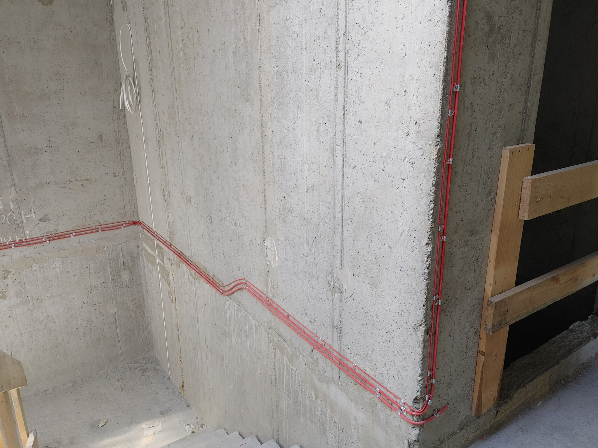Czerwone kable elektryczne prowadzone po surowej, betonowej ścianie w nowym budynku w trakcie budowy, zabezpieczone metalowymi uchwytami.