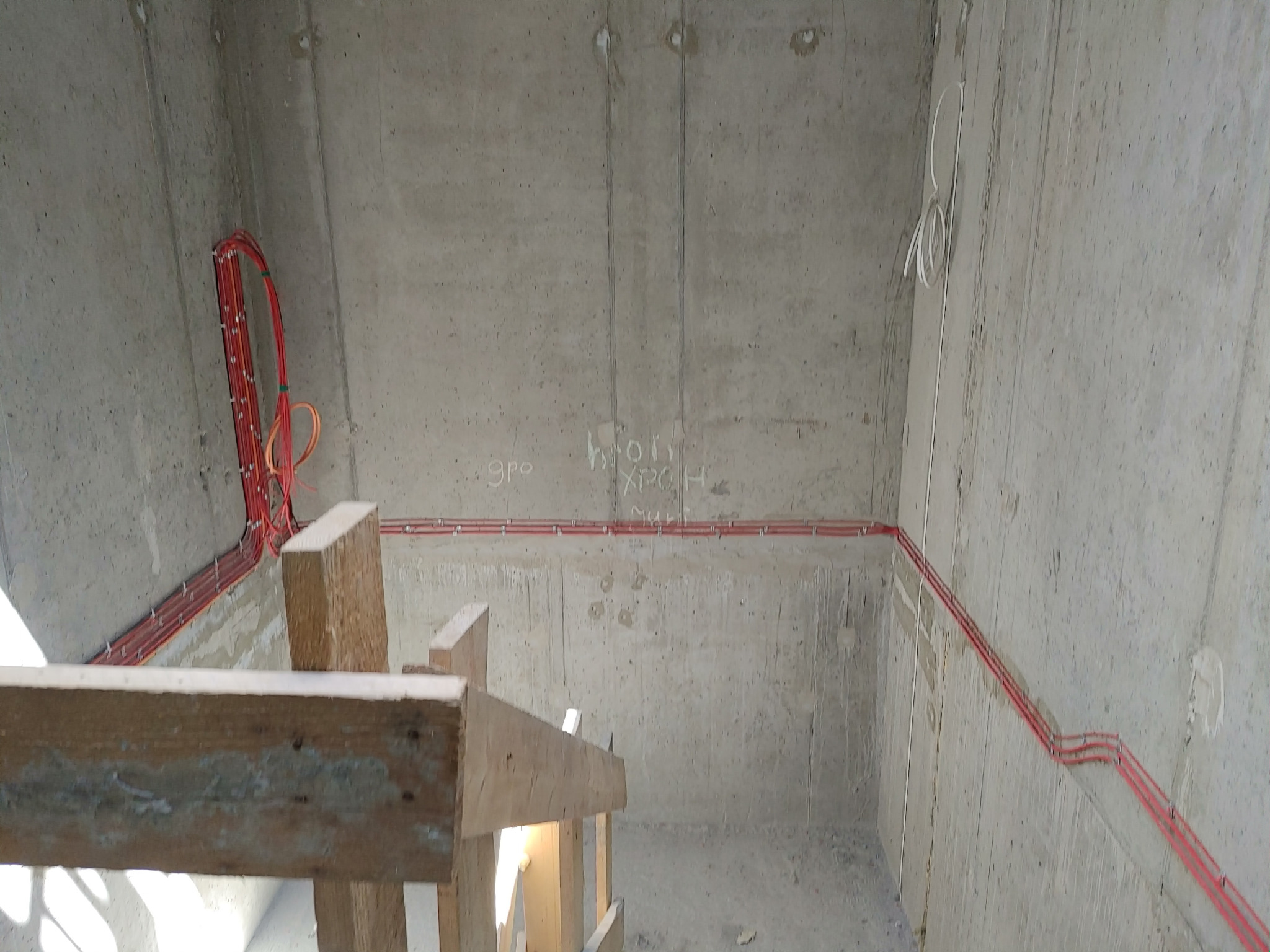 Surowy betonowy szyb windowy w trakcie budowy z widocznymi czerwonymi kablami elektrycznymi prowadzonymi wzdłuż ścian i drewnianym szalunkiem na pierwszym planie.