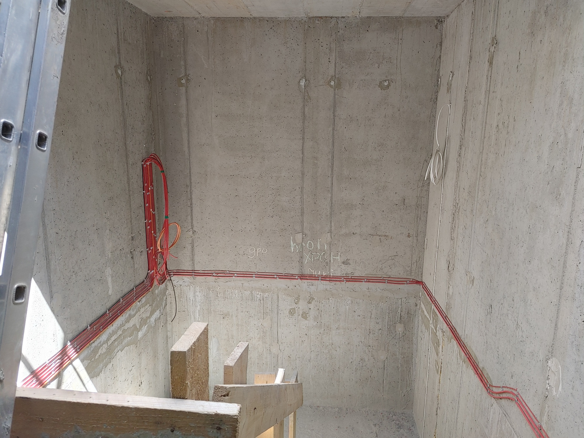Surowy betonowy szyb z częściowo ułożonymi czerwonymi kablami elektrycznymi i fragmentem drabiny aluminiowej po lewej stronie.