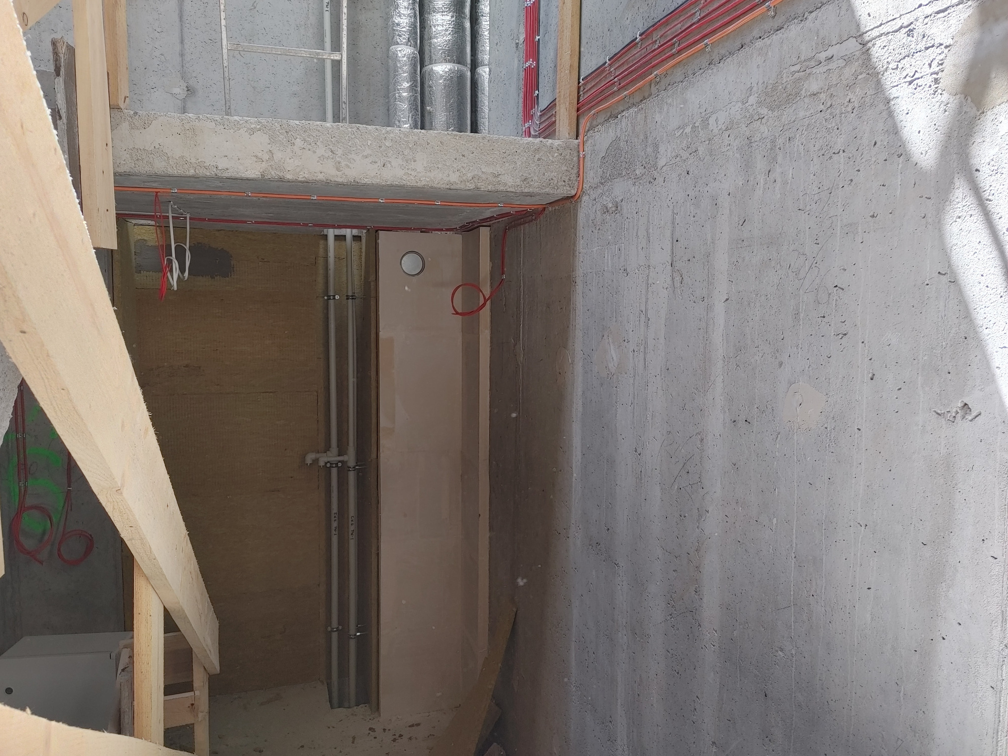 Surowy betonowy korytarz w trakcie instalacji elektrycznej: widoczne kable w peszlach, drabina, ocieplone rury wentylacyjne pod sufitem.