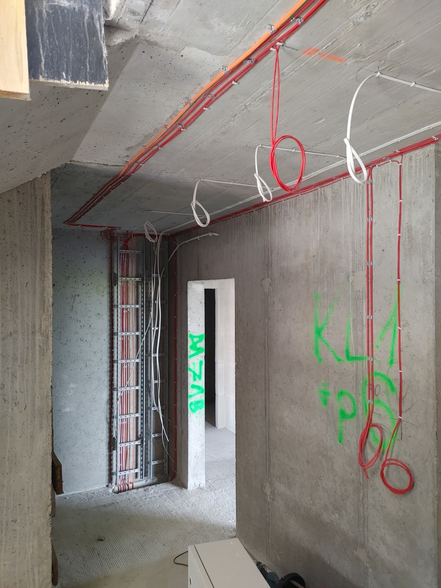 Surowy stan instalacji elektrycznej w nowym budynku w Proszowicach. Widoczne kable prowadzone po ścianach i suficie, częściowo w korytach kablowych, z zielonymi napisami na ścianie.