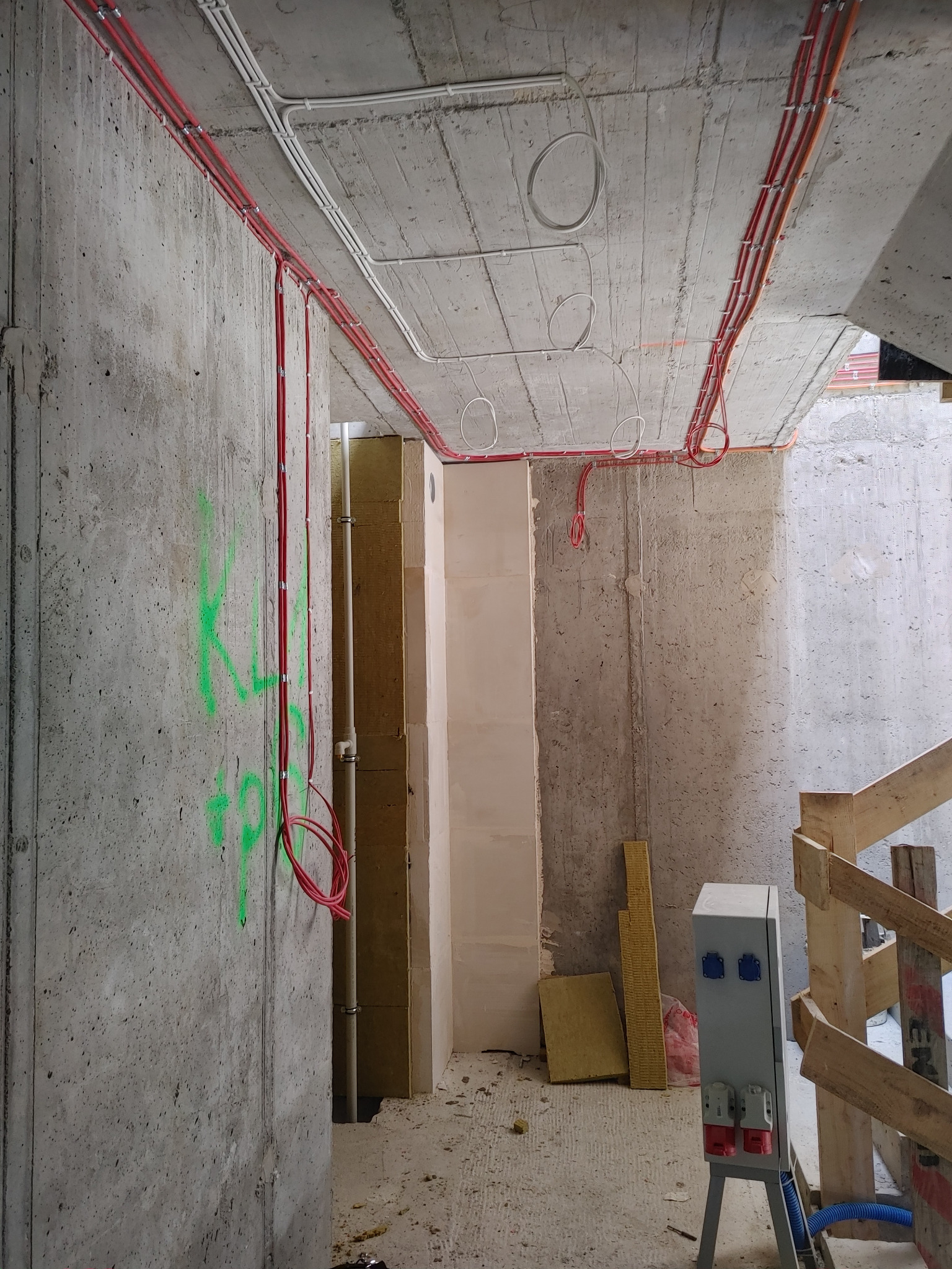 Surowy betonowy strop z widocznymi czerwonymi i białymi rurami instalacji elektrycznej, szara skrzynka elektryczna na stojaku, ściany z zielonymi napisami, izolacja termiczna w tle, budowa...